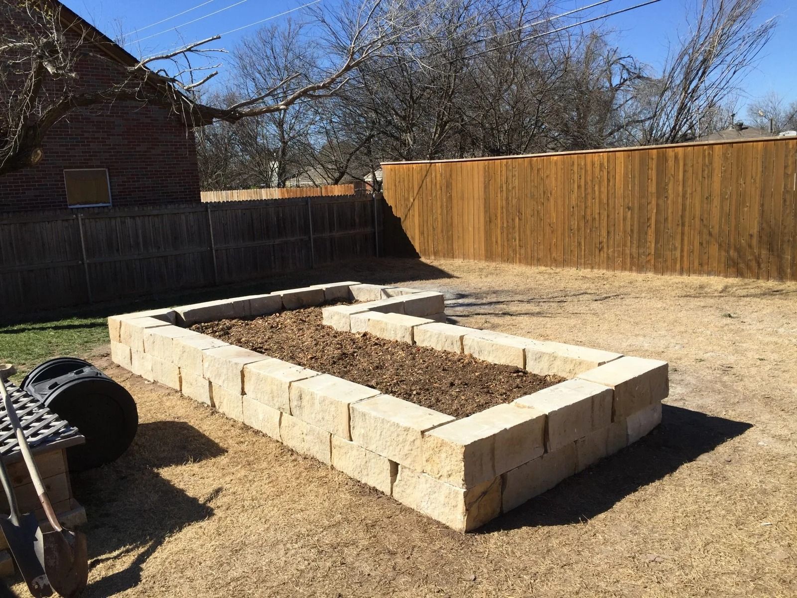 Raised stone garden bed in a sunny backyard with a wooden fence and bare trees.