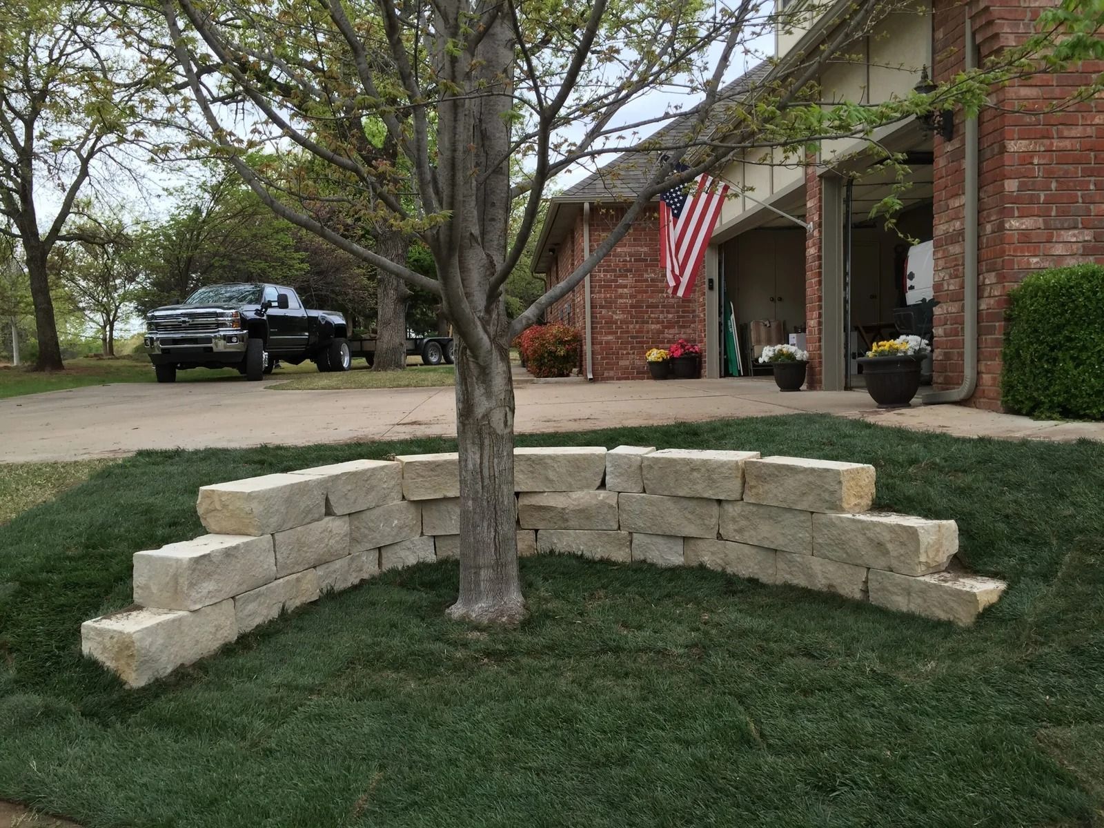 Curved stone bench around a tree in a front yard beside a brick house and American flag.