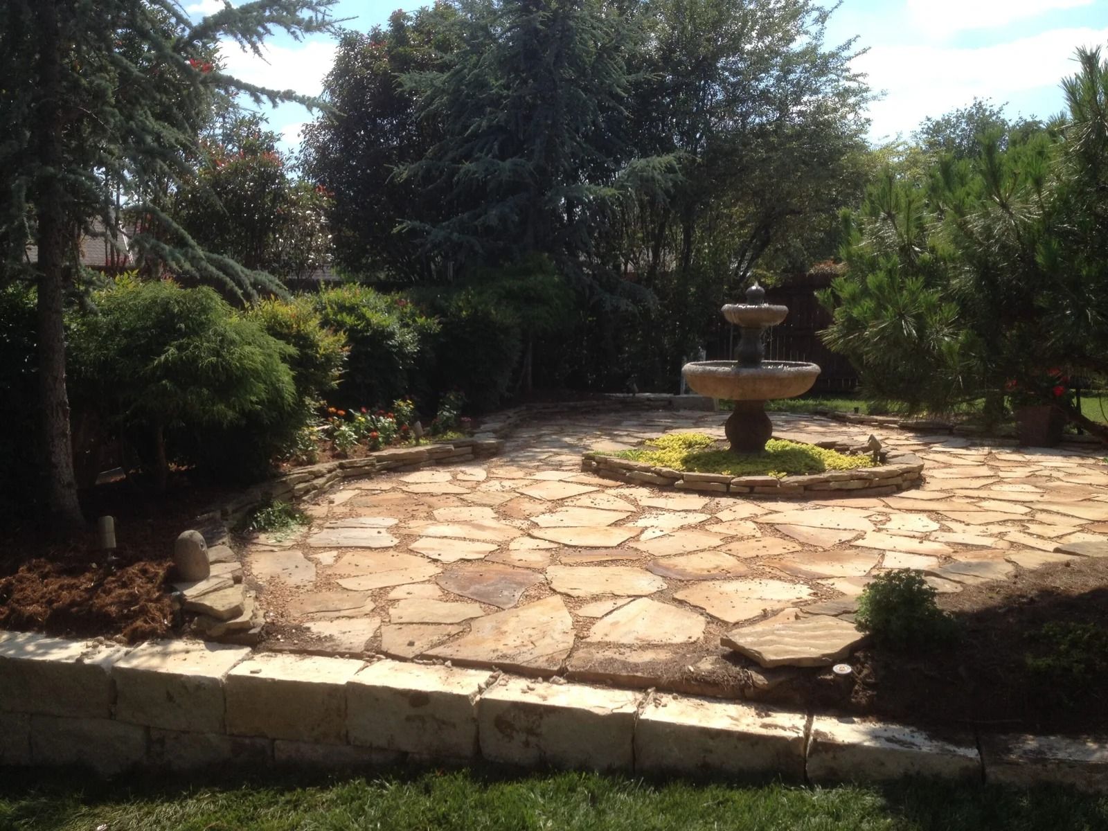 Stone patio with a small tiered fountain surrounded by lush trees and greenery in a garden.