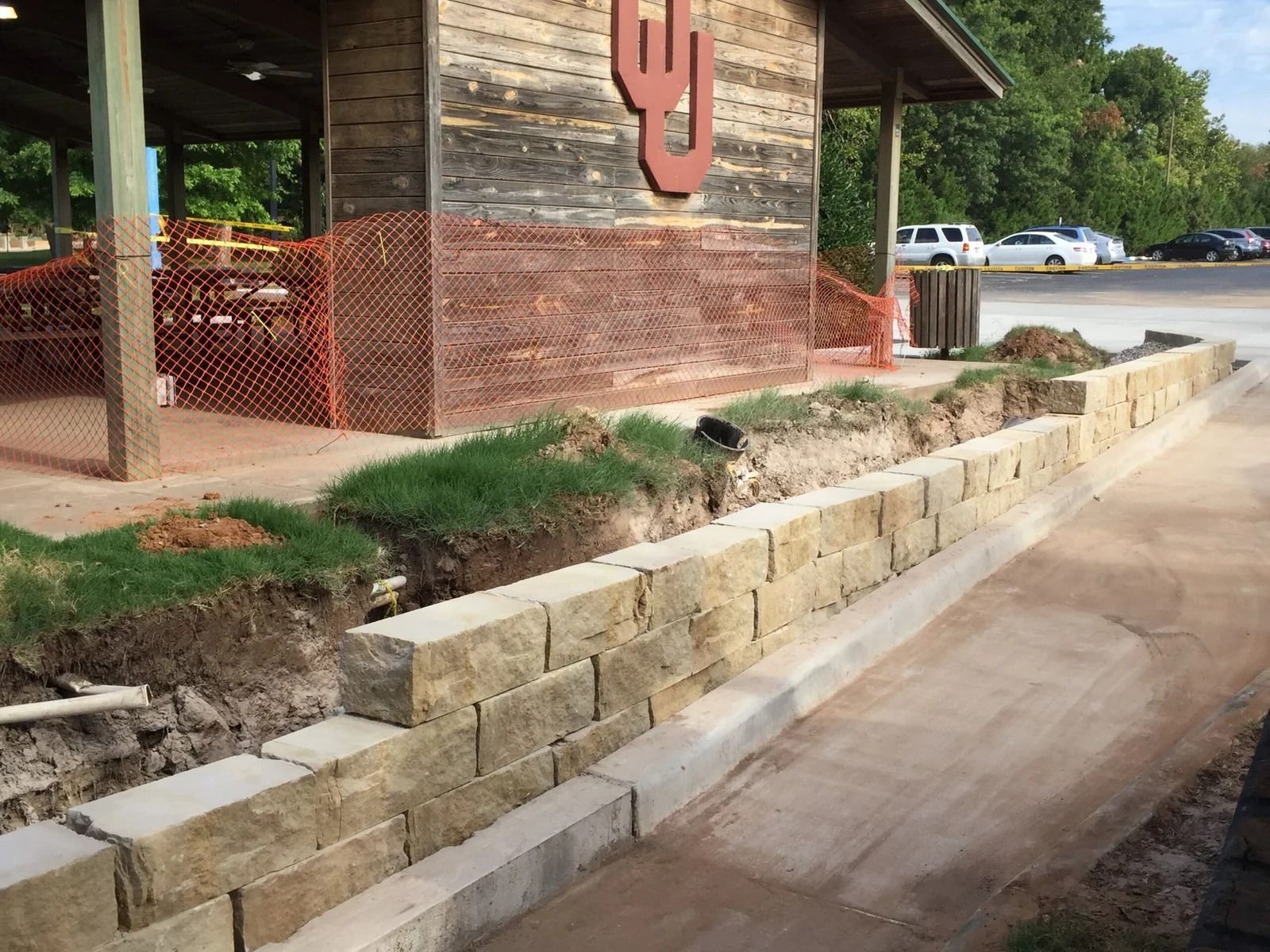Stone retaining wall beside a building walkway, with exposed soil and orange safety fencing.