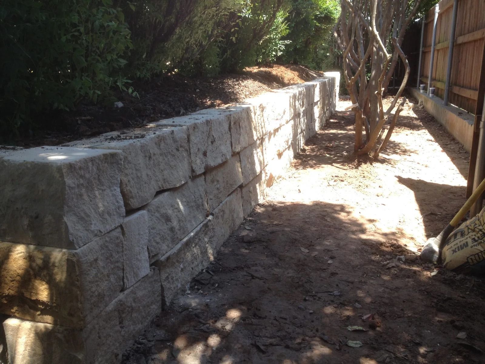 Stone retaining wall along a narrow dirt path with trees and sunlight in a garden area