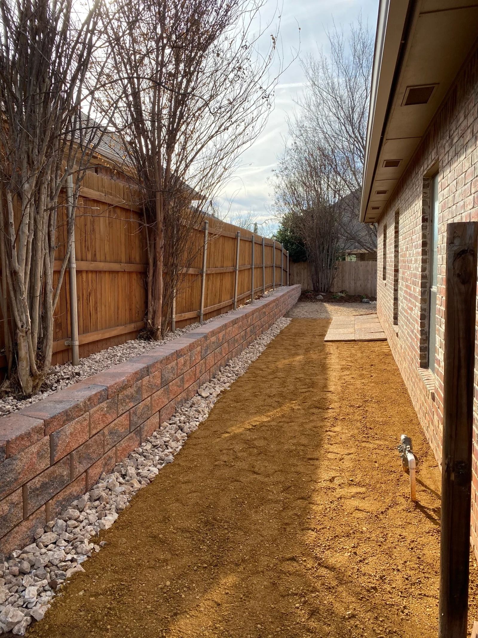 Narrow backyard with wooden fence, stone border, and dry grass beside a house.