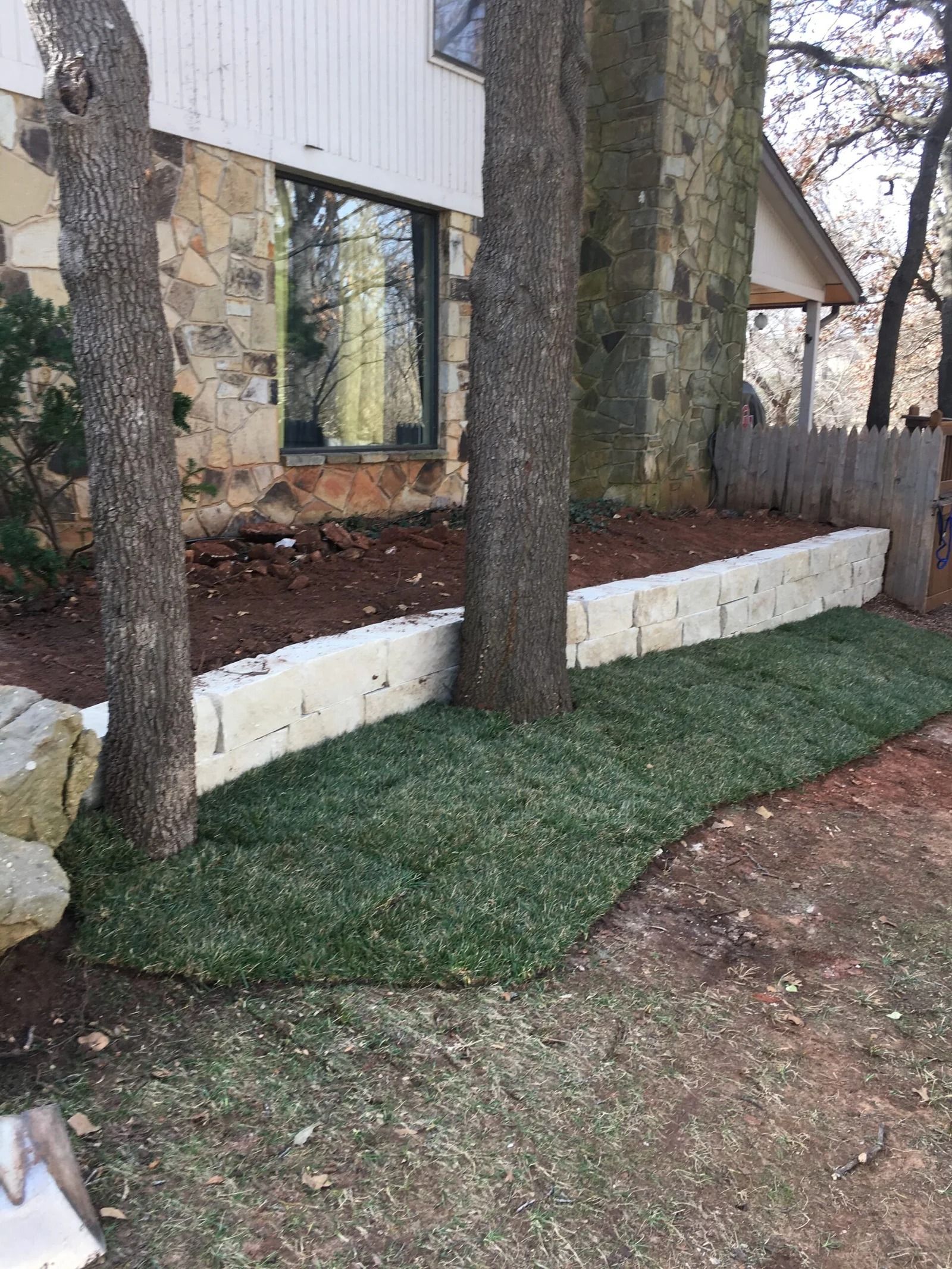 House exterior with stone trees, low white retaining wall, and mulch bed beside a green lawn.