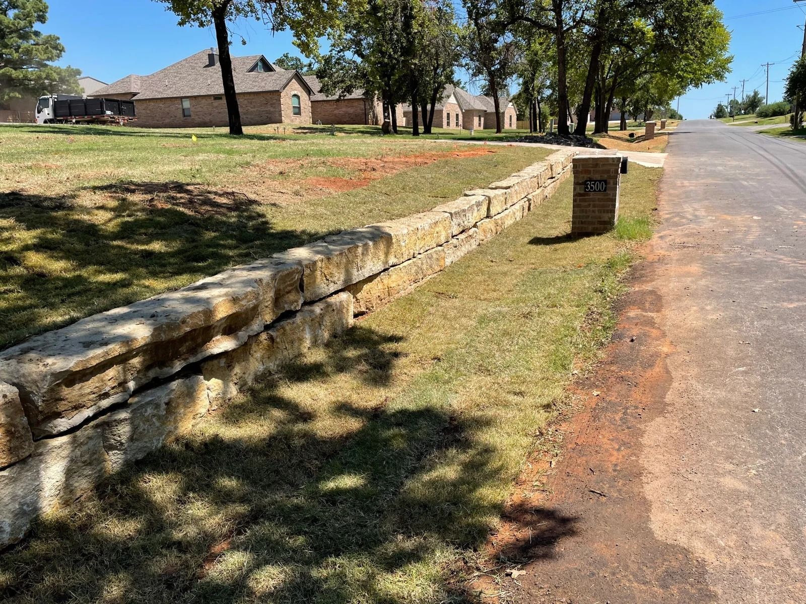Stone retaining wall beside a quiet road, with trees, a grassy verge, and a house in the background