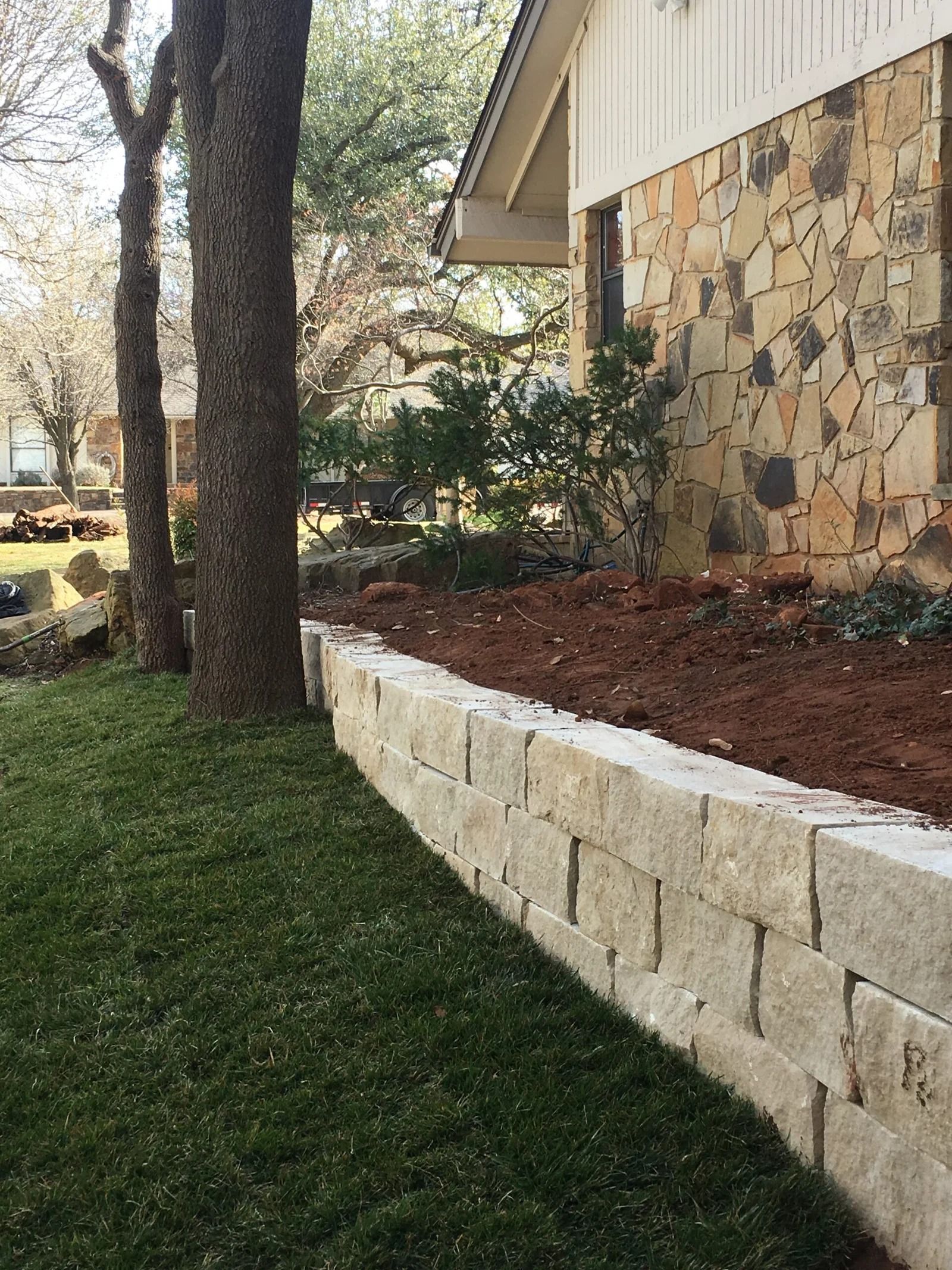 Stone retaining wall beside a grassy yard and trees next to a house with stone siding