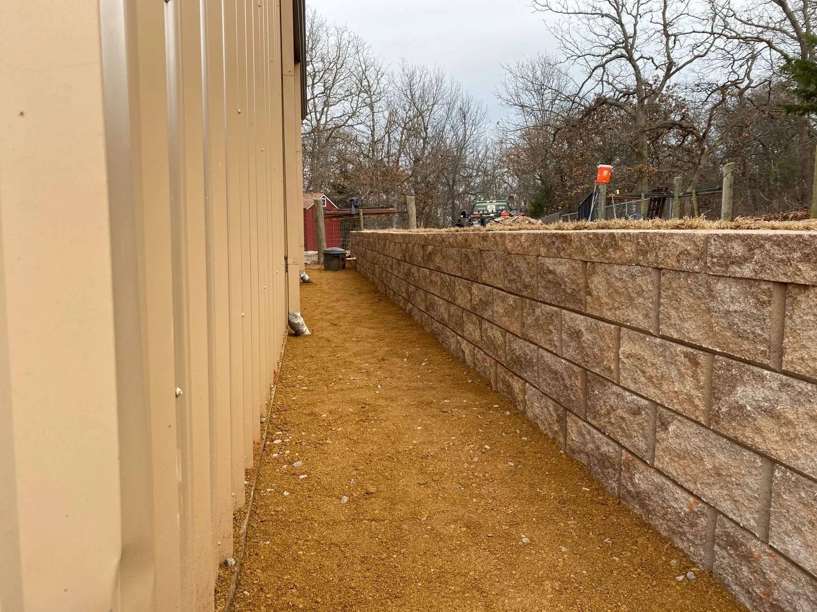 Narrow gravel path between a tan building wall and a stone retaining wall, lined with bare trees.