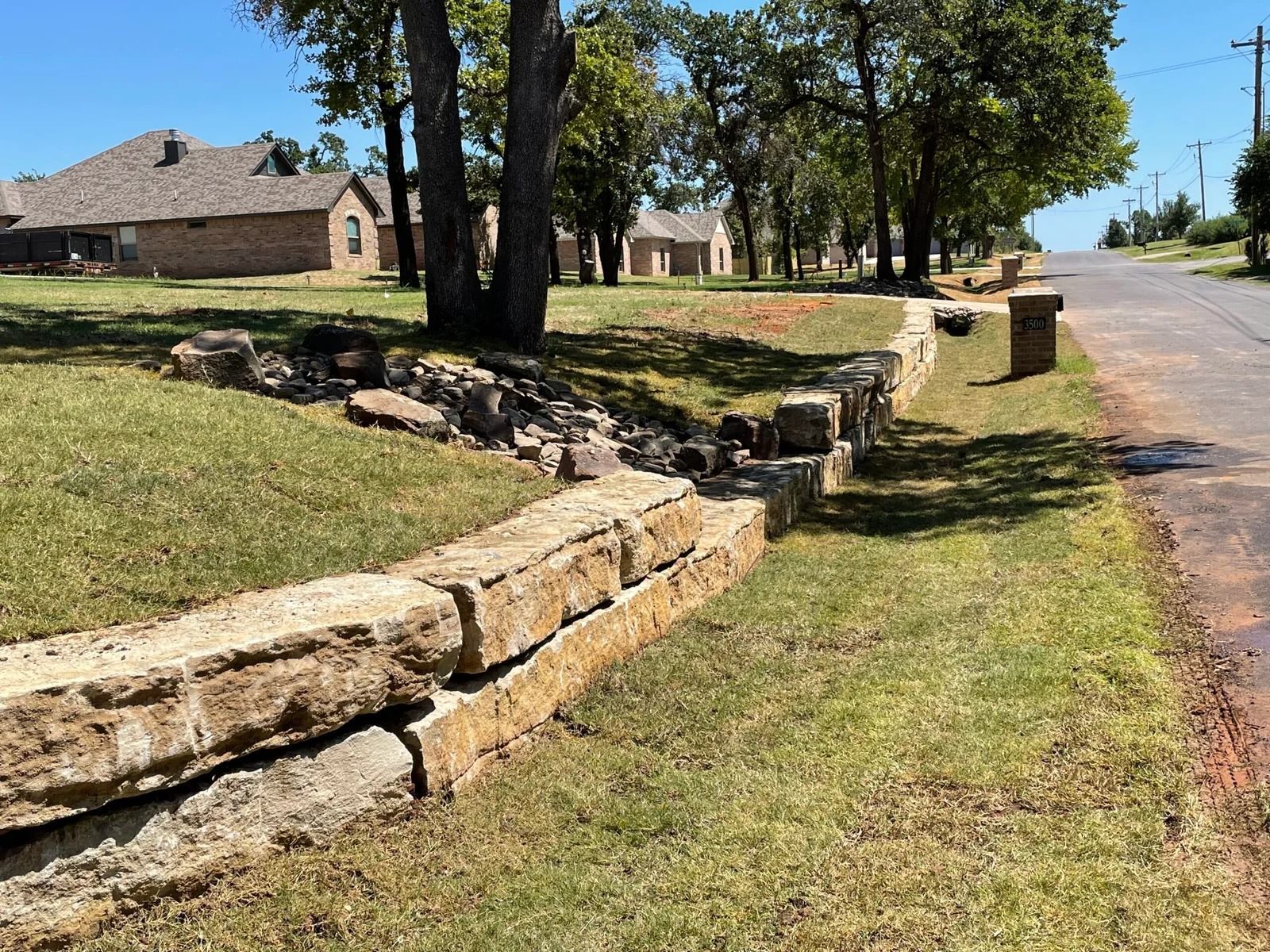 Stone retaining wall along a grassy roadside beside a tree-lined residential street