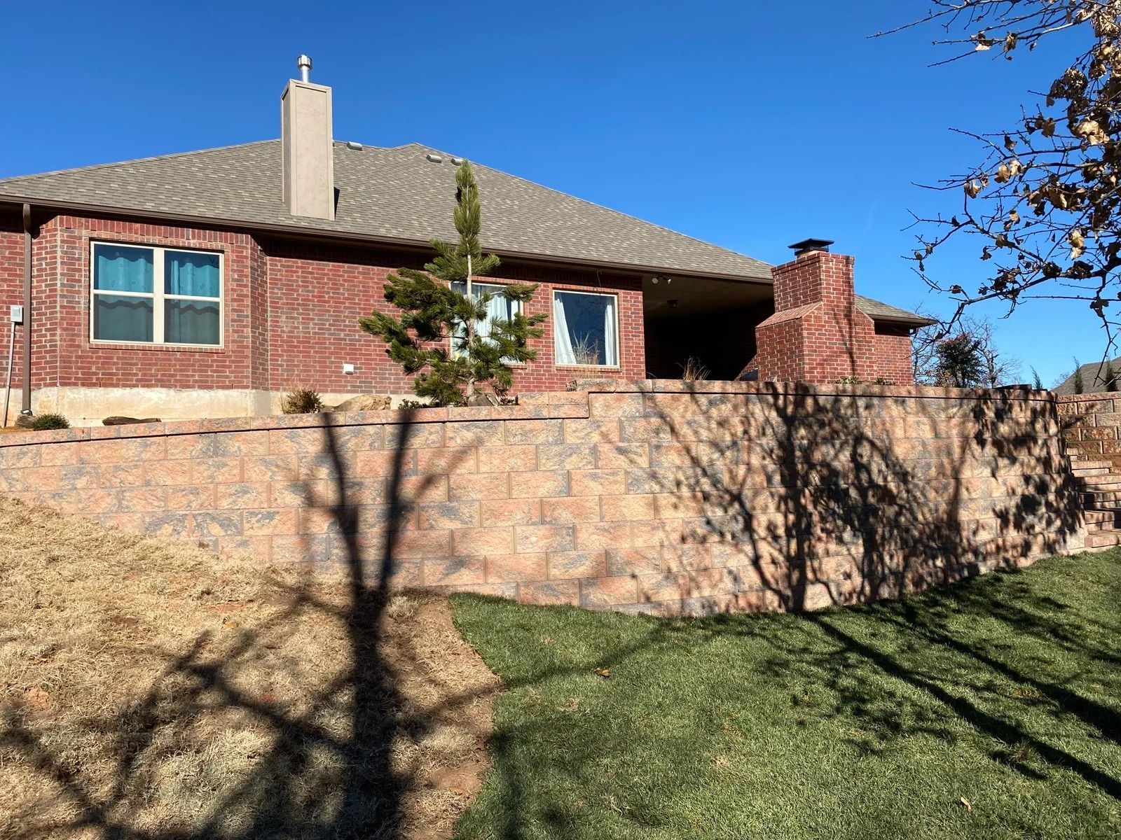 Brick house with tan stone wall, red-tiled roof, and grassy yard under a clear blue sky