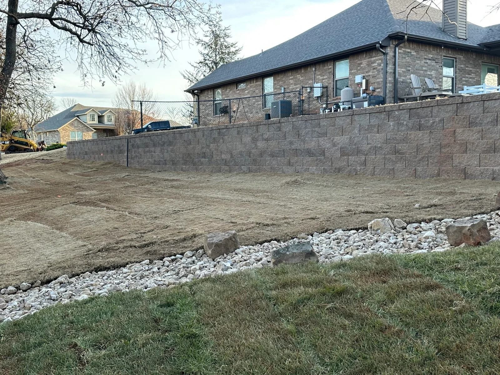 Bare dirt lot beside a stone house and retaining wall, with grass in the foreground.