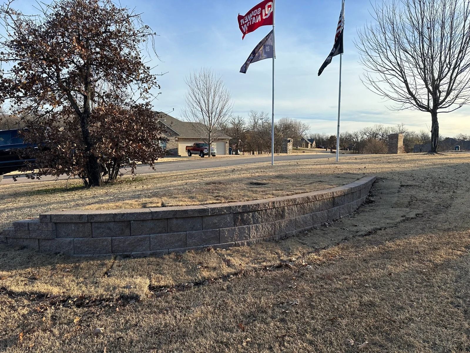 Grassy roadside median with two flags, a tree, and a low stone retaining wall on a clear day