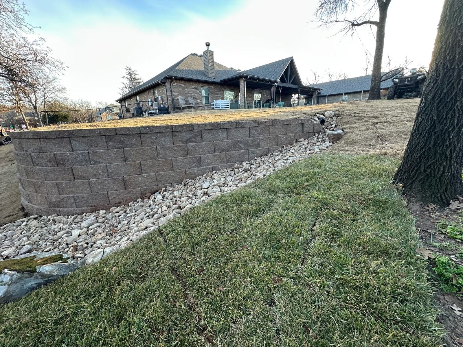 Stone retaining wall beside a grassy slope, with a house and trees in the background.