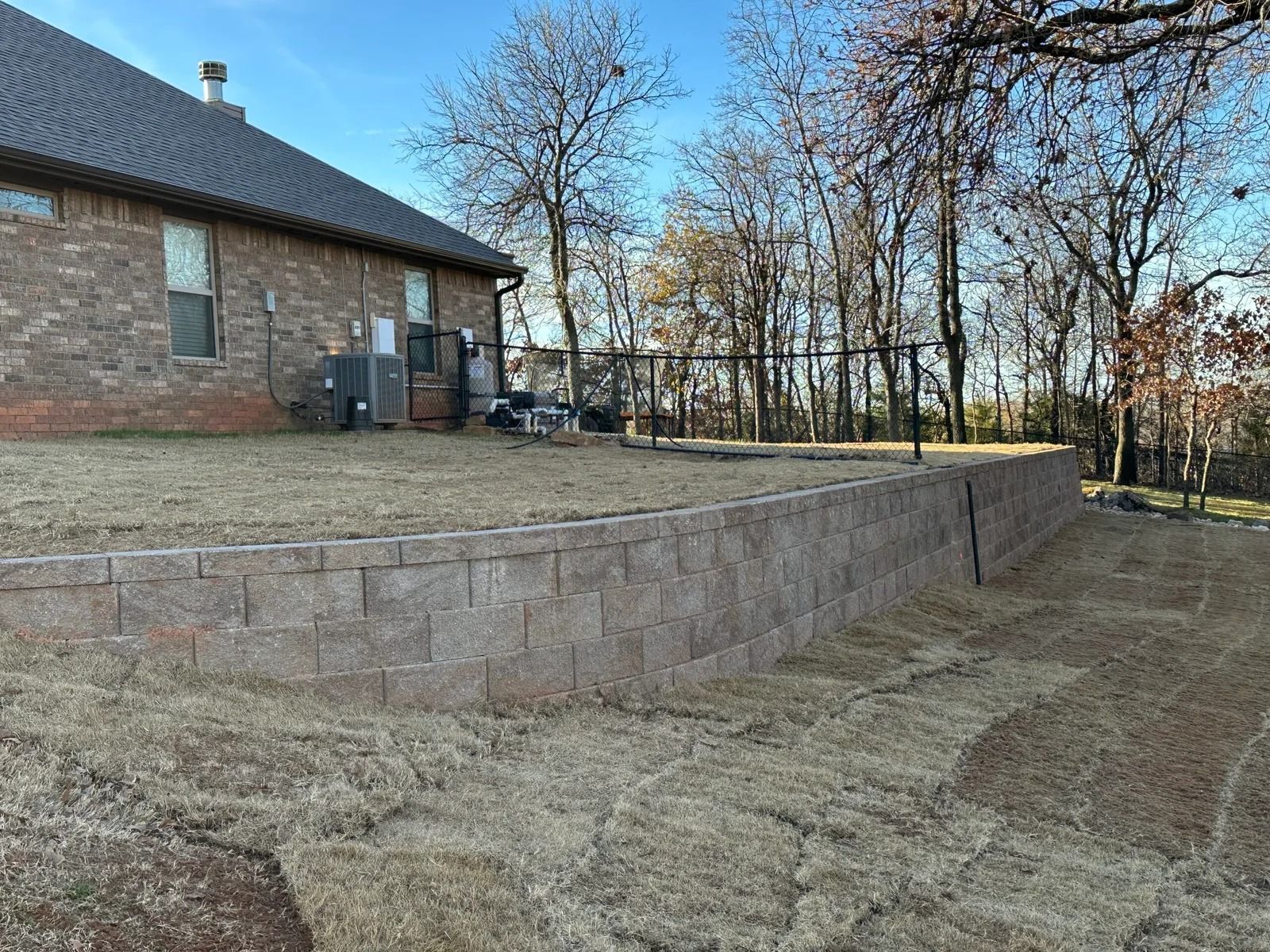Brick house with a raised stone retaining wall and a bare backyard overlooking leafless trees.