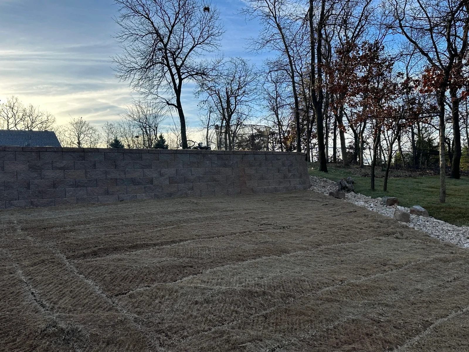 Bare dirt lot beside a low stone wall and leafless trees at dusk