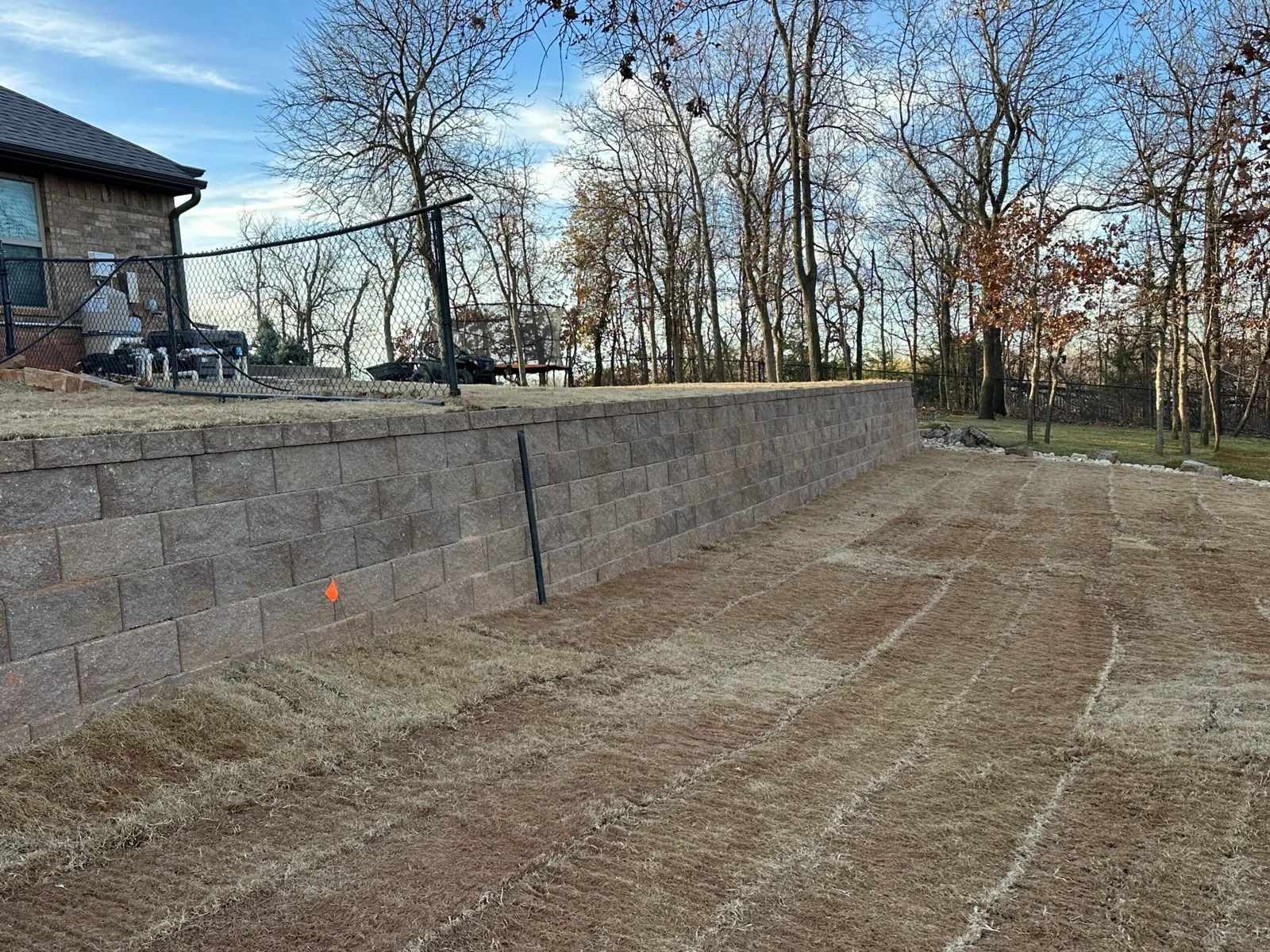 Backyard with a stone retaining wall, bare trees, and a dirt yard beside a house