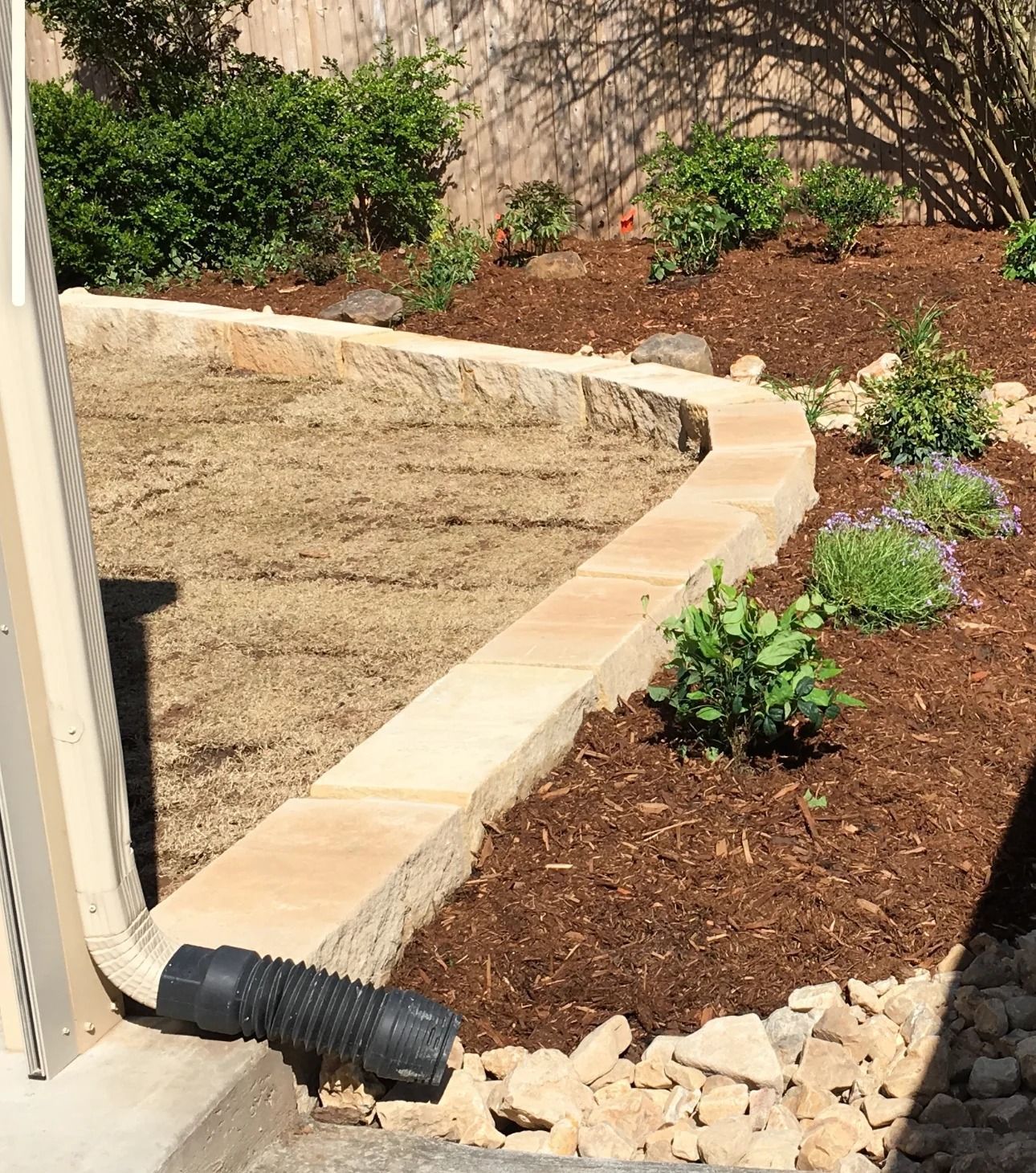 Curved tan retaining wall edging a mulched garden bed beside a lawn and downspout.