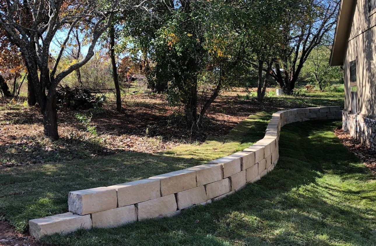 Curved tan block retaining wall along a grassy yard beside trees and a house in sunlight