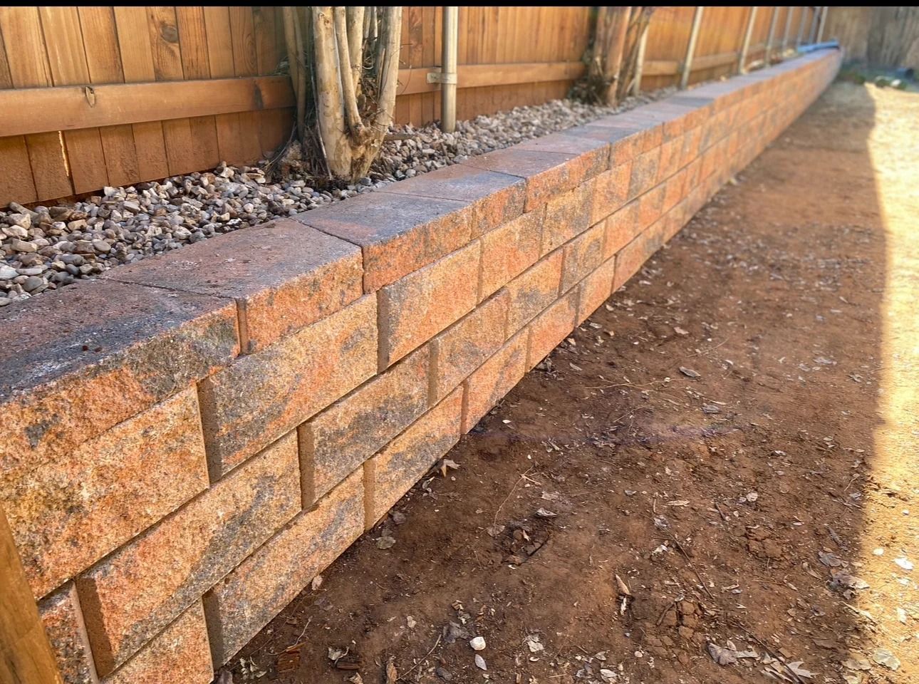 Brick retaining wall bordering a dirt path beside a wooden fence and gravel bed