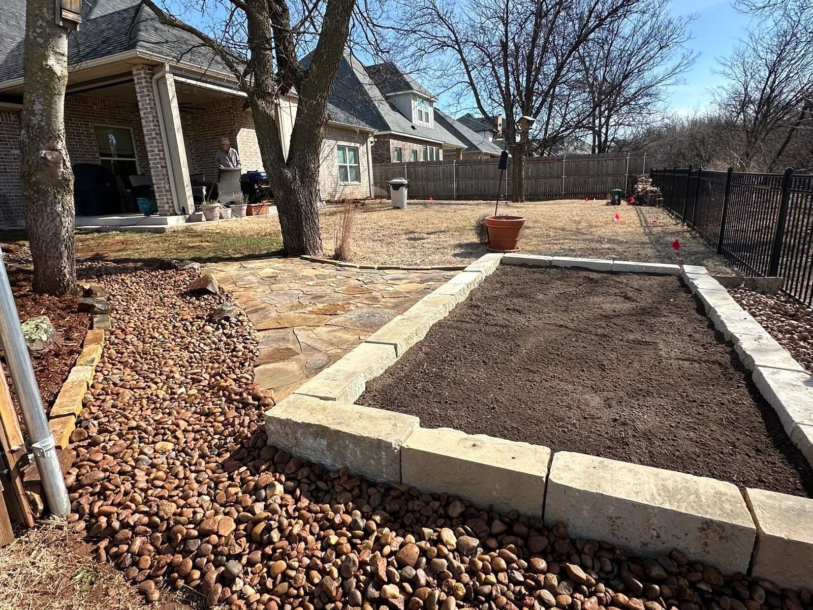 Backyard with raised garden bed, bare soil, and stone border beside a house and patio.