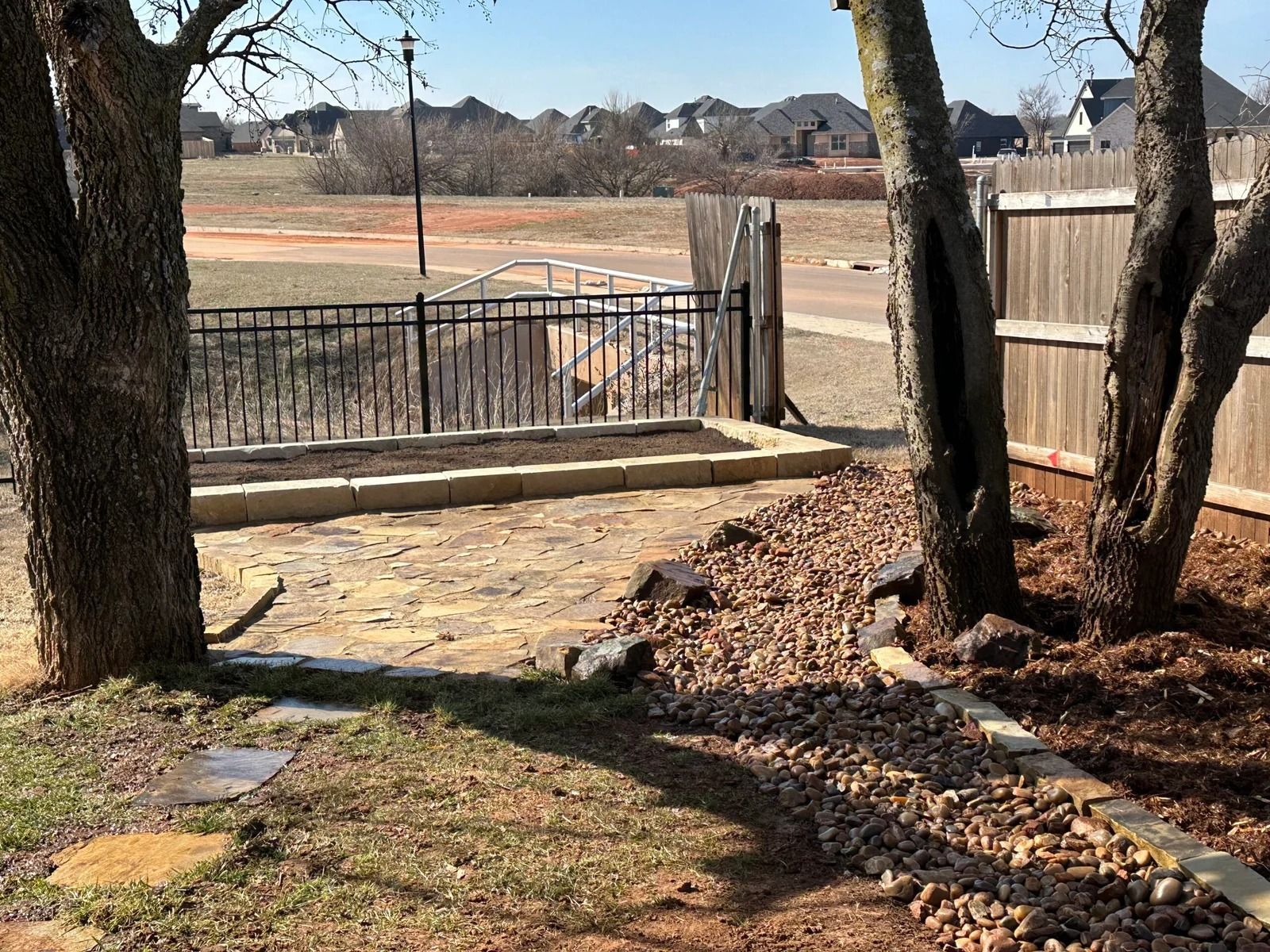 Backyard patio with stone path, trees, wooden fence, and black gate overlooking a road under clear sky