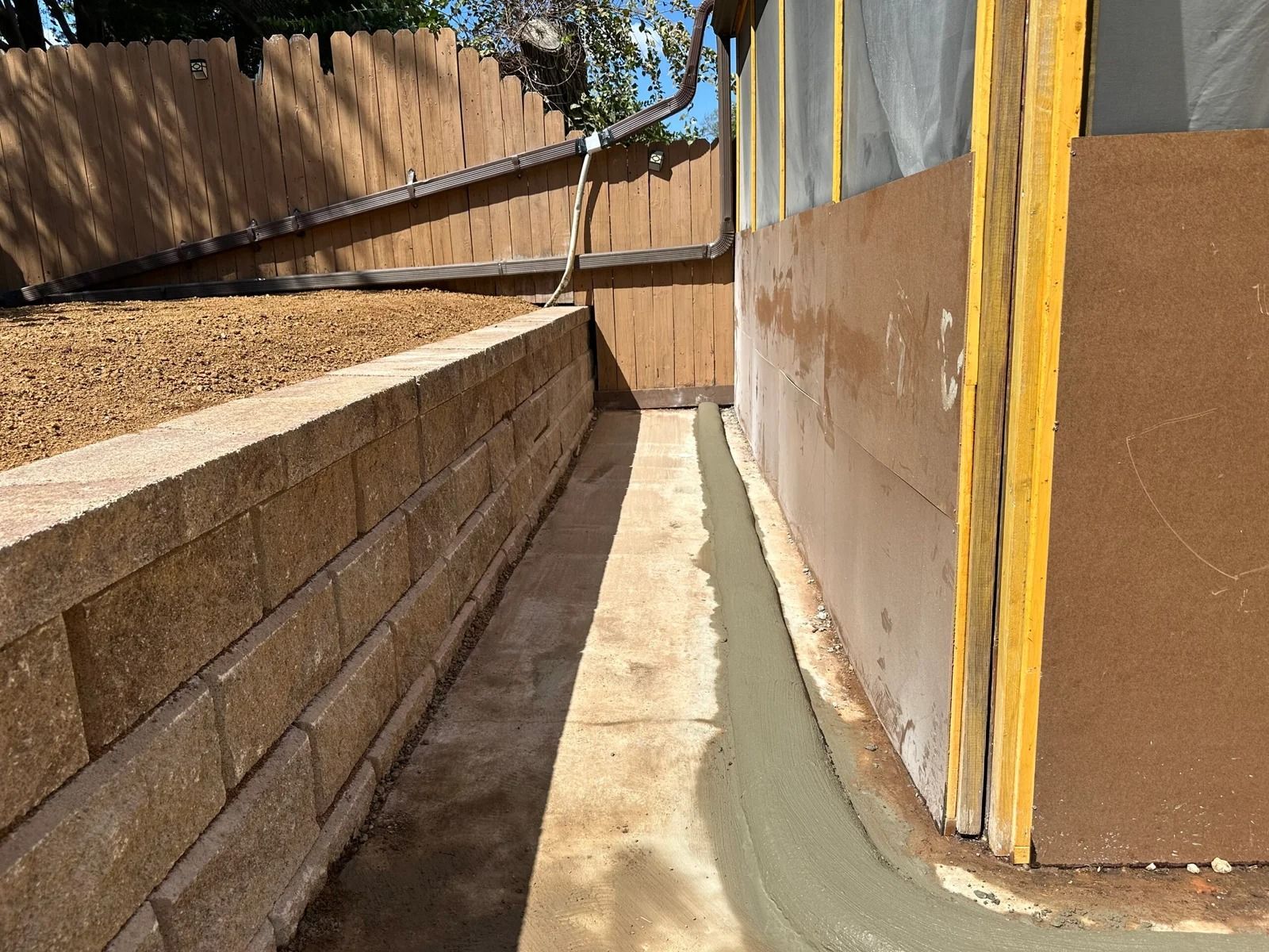 Narrow concrete drainage channel beside a tan retaining wall and wooden fence in a backyard
