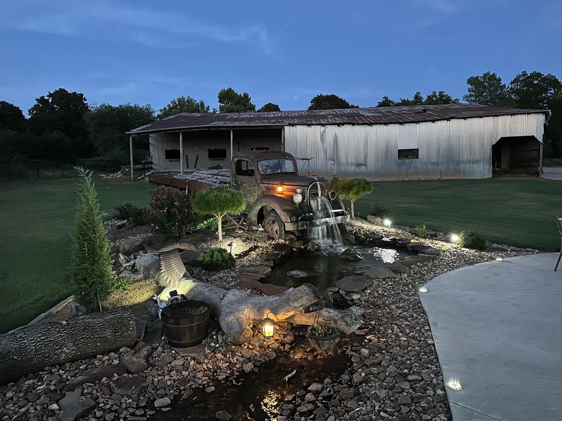 Lit backyard pond and waterfall with rocks, plants, and a house in the background at dusk