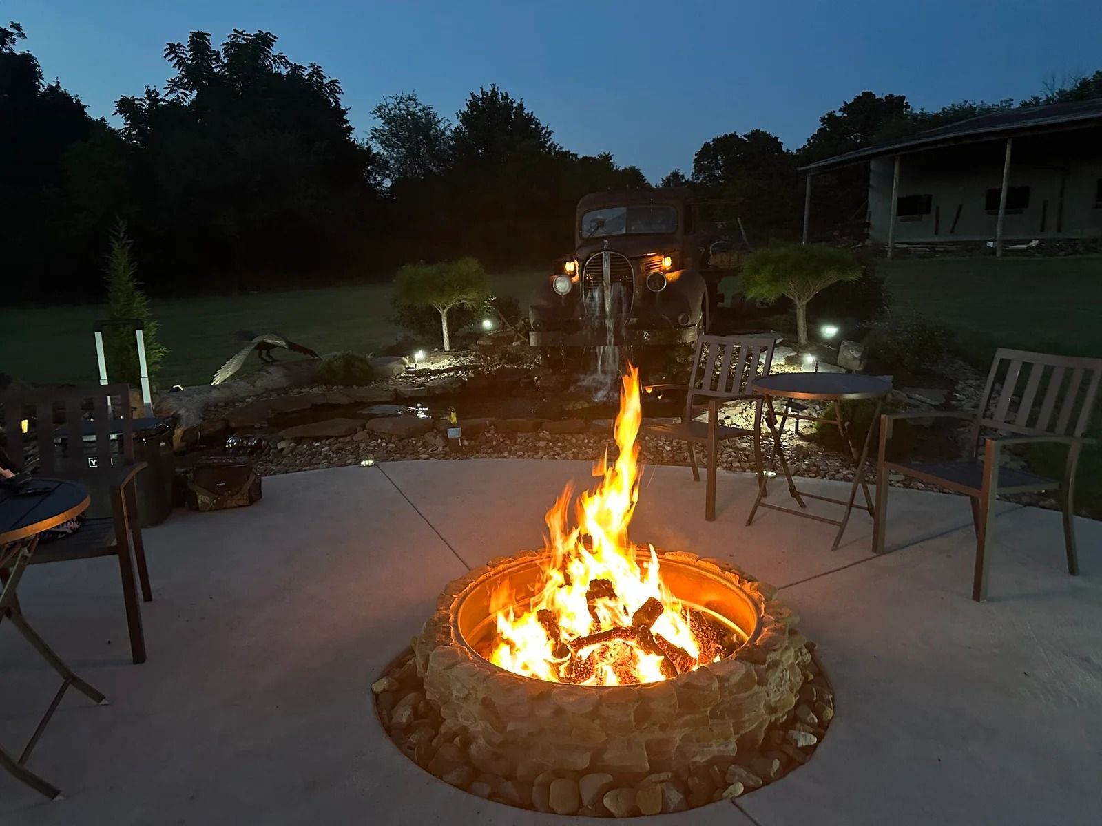 Backyard patio at dusk with a lit stone fire pit and outdoor chairs