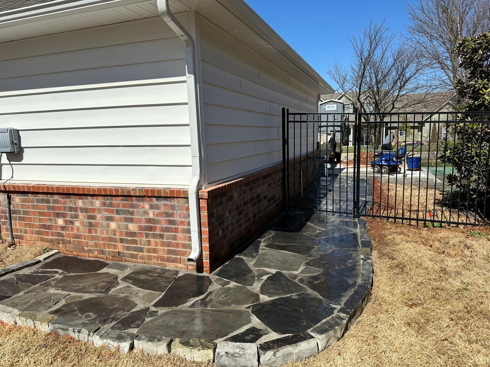Stone walkway beside a brick-and-siding house leads to a black metal gate in a fenced yard.