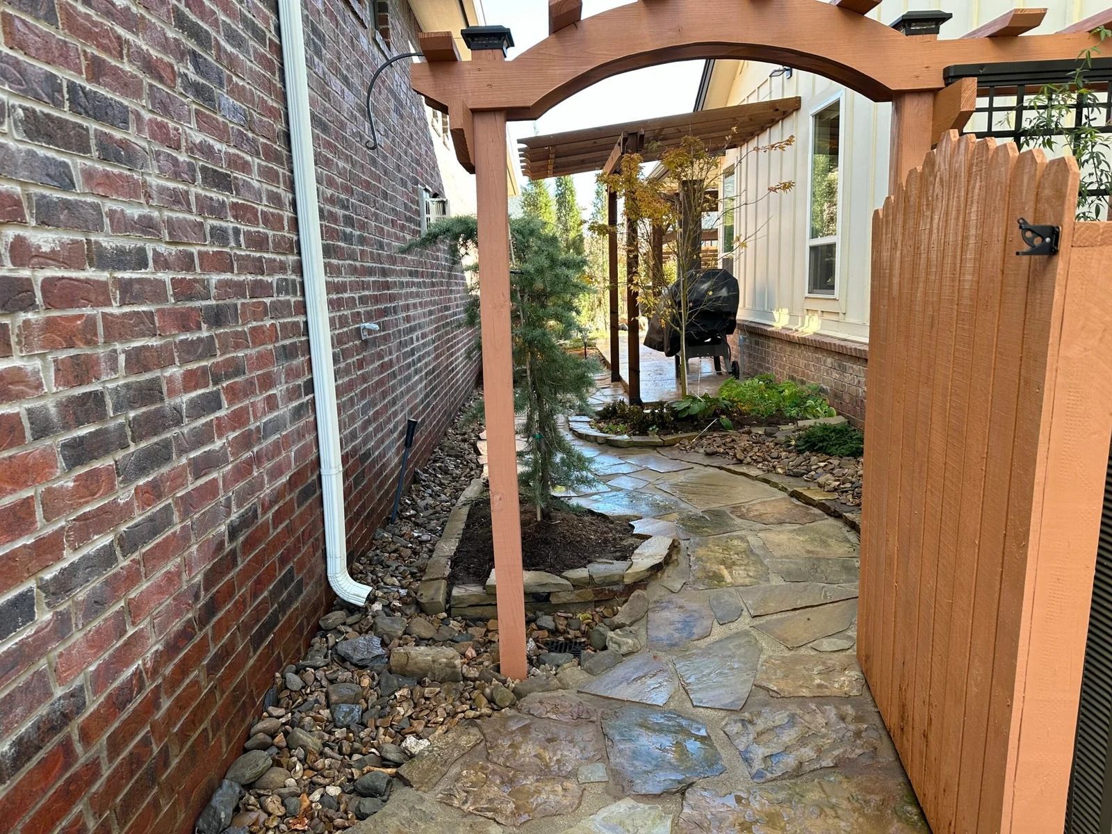 Narrow stone walkway beside brick wall and house, with wooden archway and fence gate.