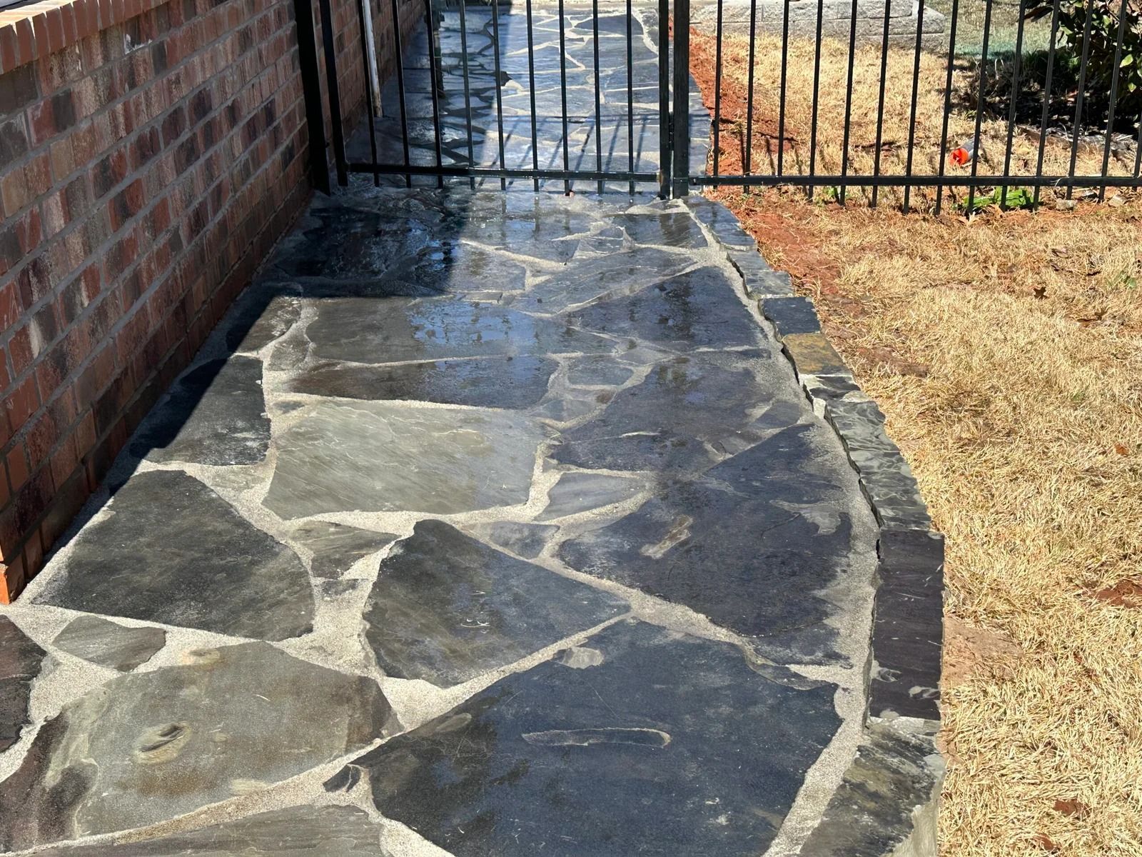 Stone walkway beside a brick wall and black gate, bordered by dry grass.
