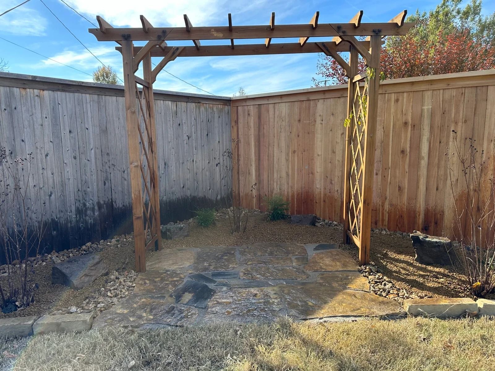 Wooden pergola in a small fenced backyard with a stone patio and clear blue sky