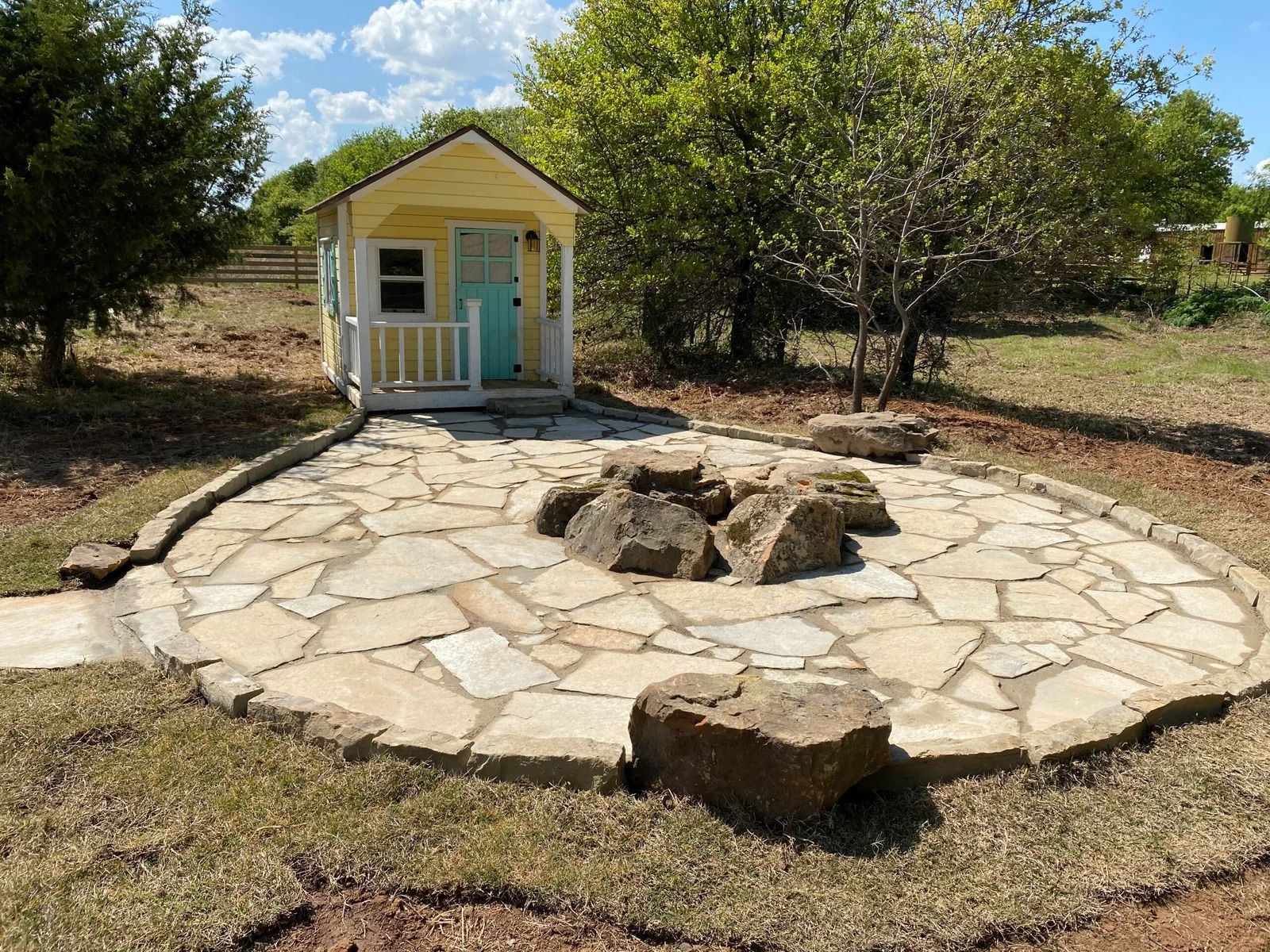 Small yellow garden shed beside a circular stone patio with rocks and trees in a sunny yard