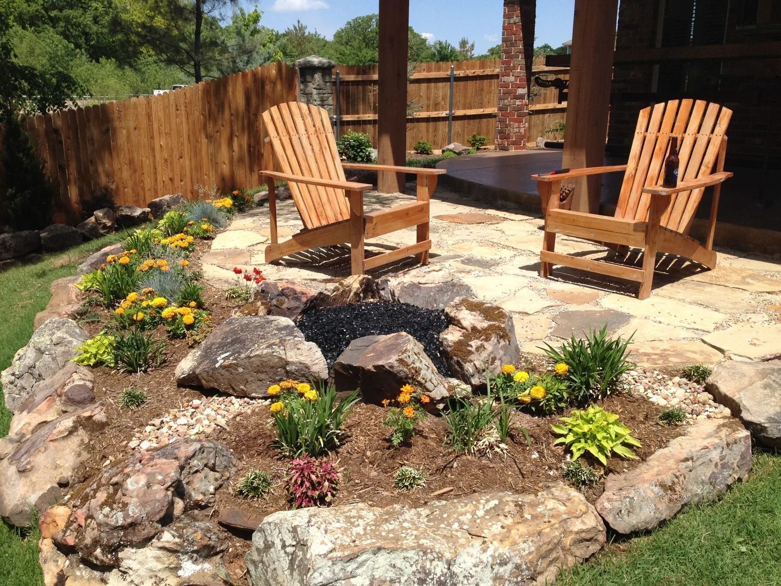 Wooden lounge chairs beside a rock fire pit in a landscaped backyard patio