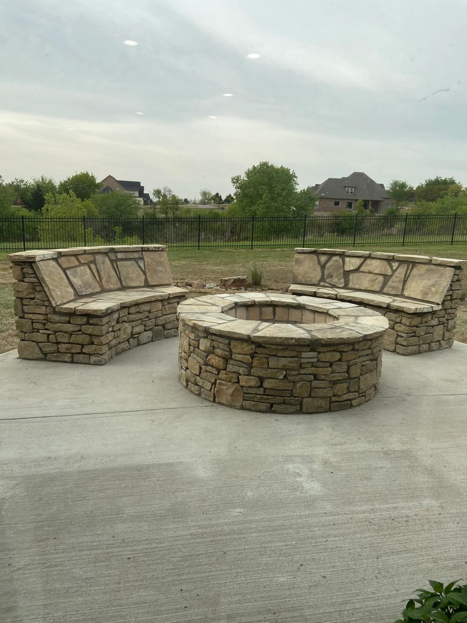 Stone fire pit with circular seating benches in a backyard patio with grassy field beyond