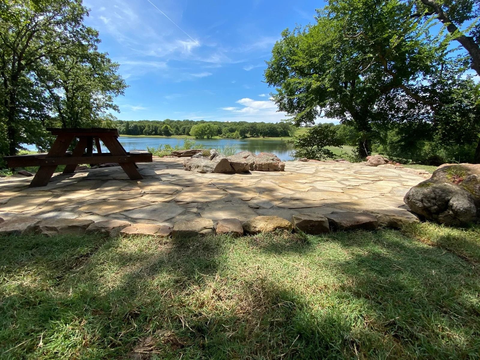 Shaded lakeside picnic area with a stone patio, picnic table, trees, and calm water beyond.