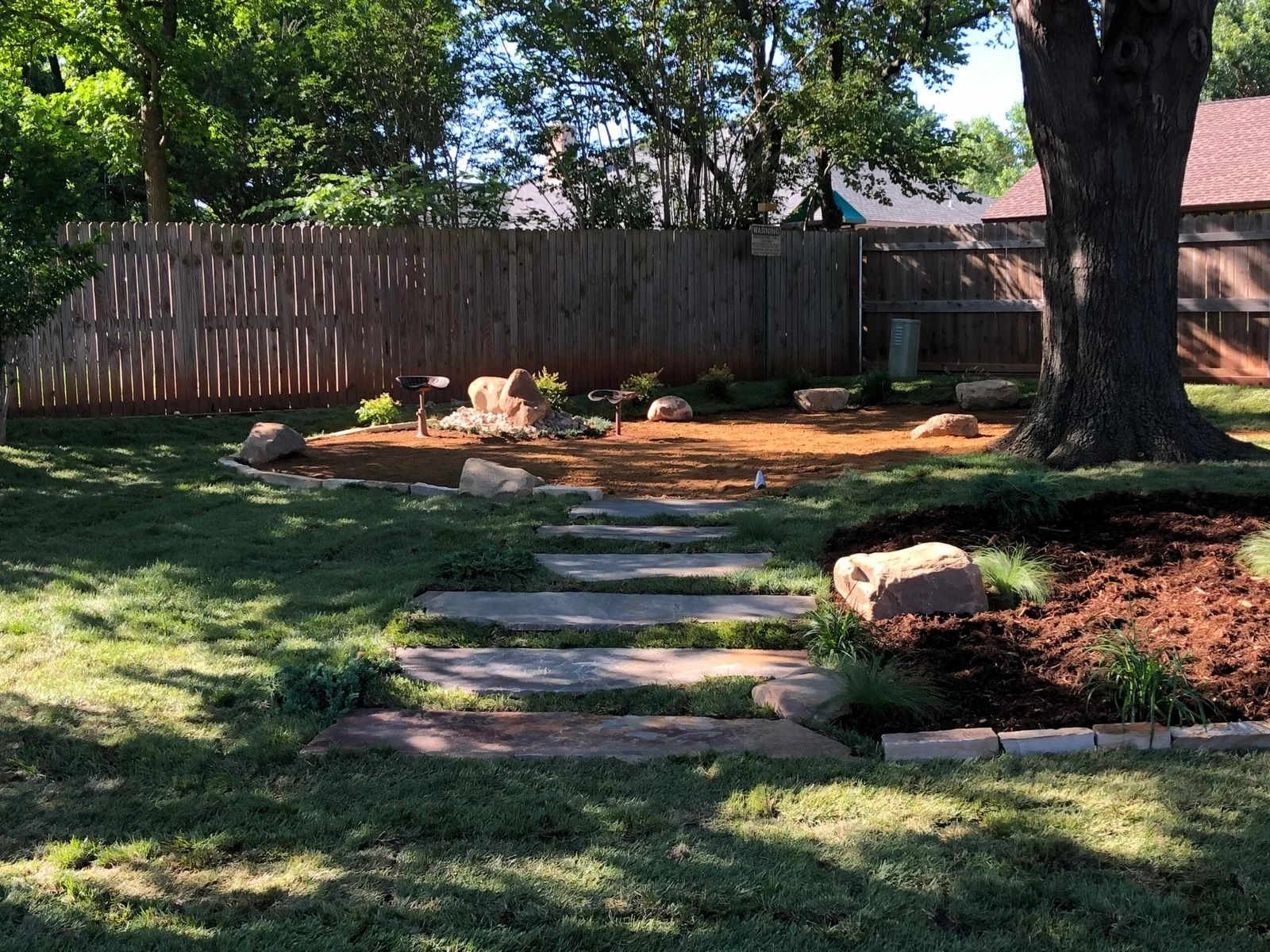 Backyard with stone path, grass, trees, wooden fence, and a mulch bed in sunlight