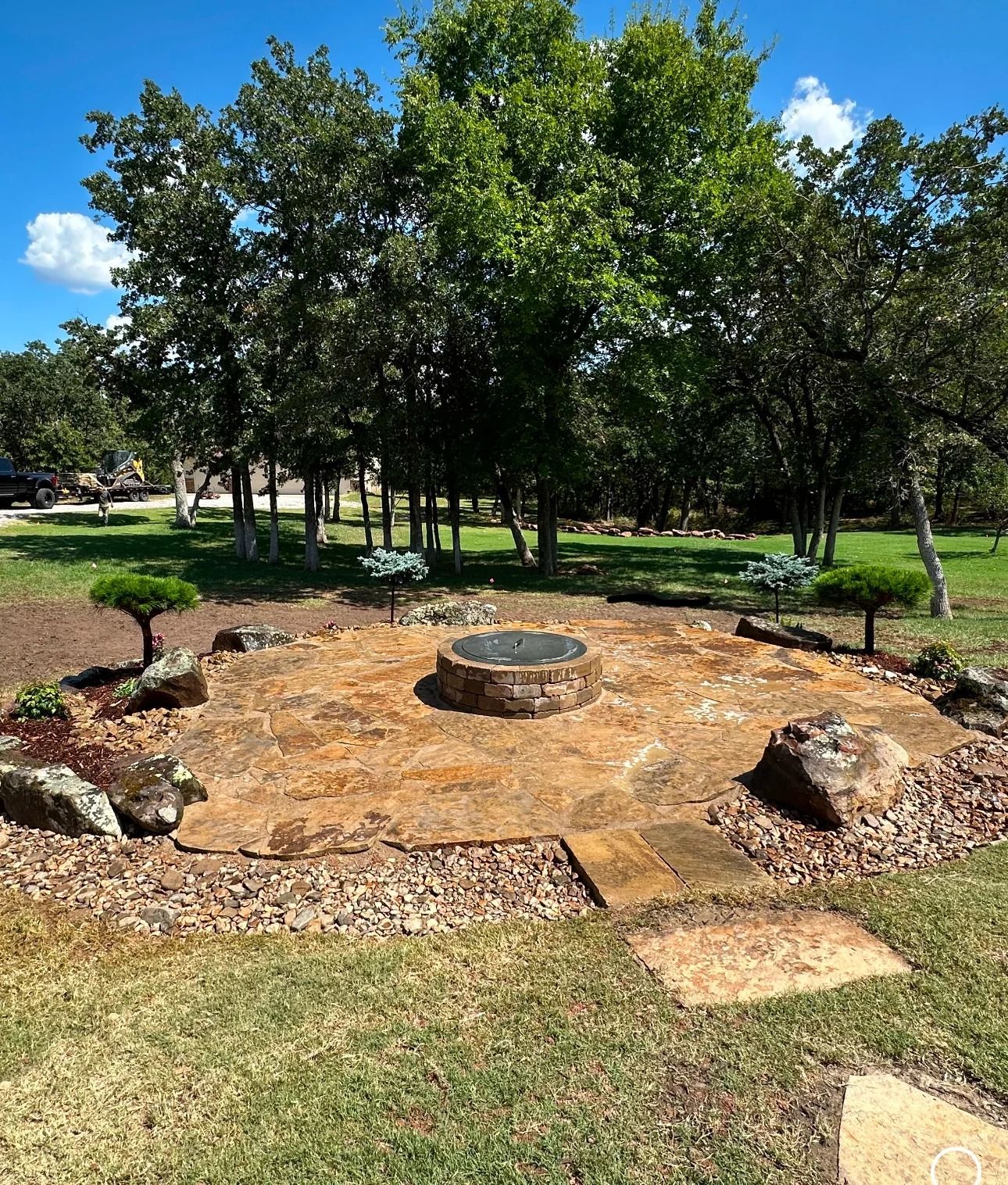 Landscaped garden with a circular stone feature, rocks, and trees under a bright blue sky
