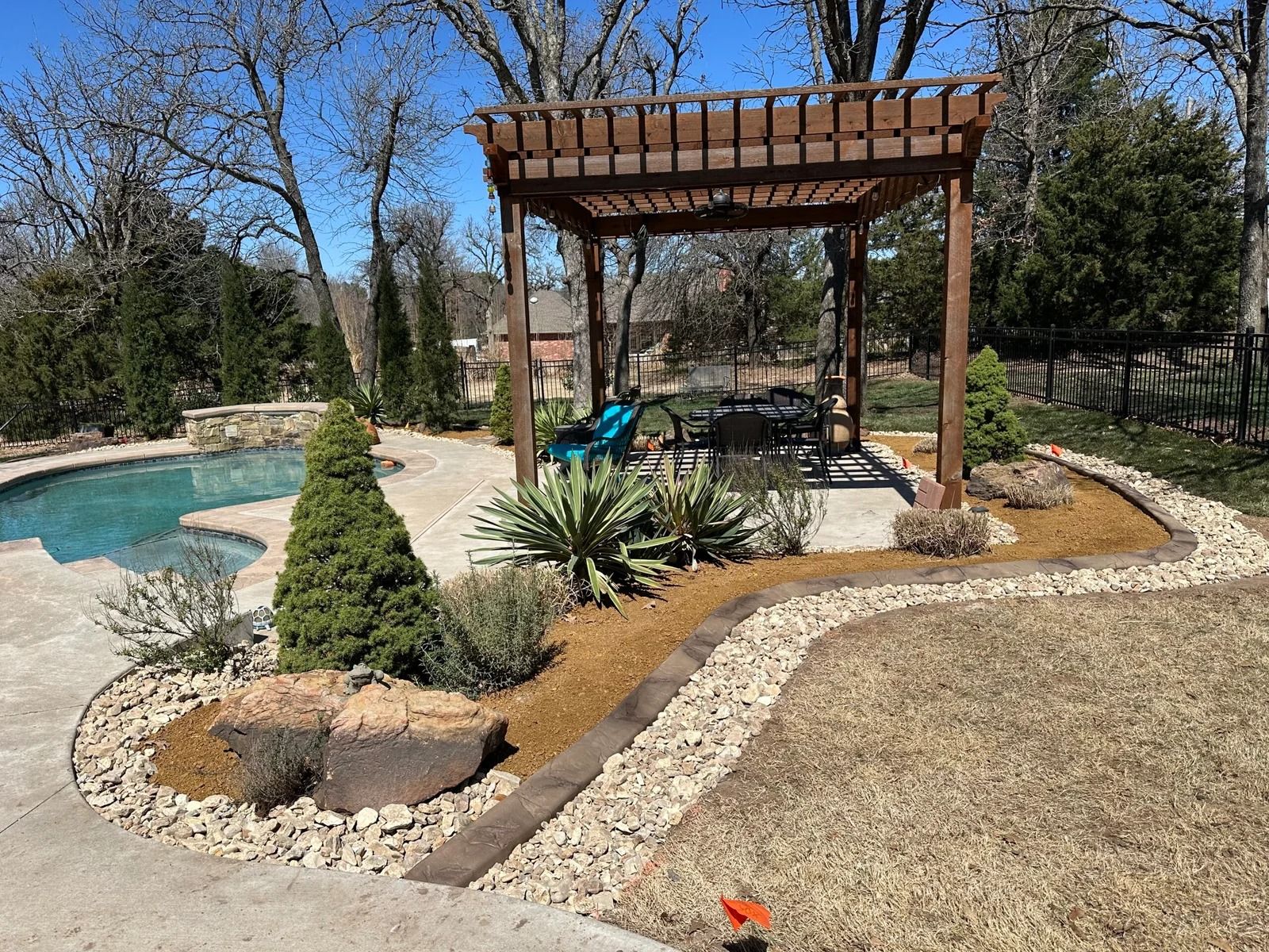 Backyard pool with pergola seating area, stone path, and landscaped garden beds under bare trees