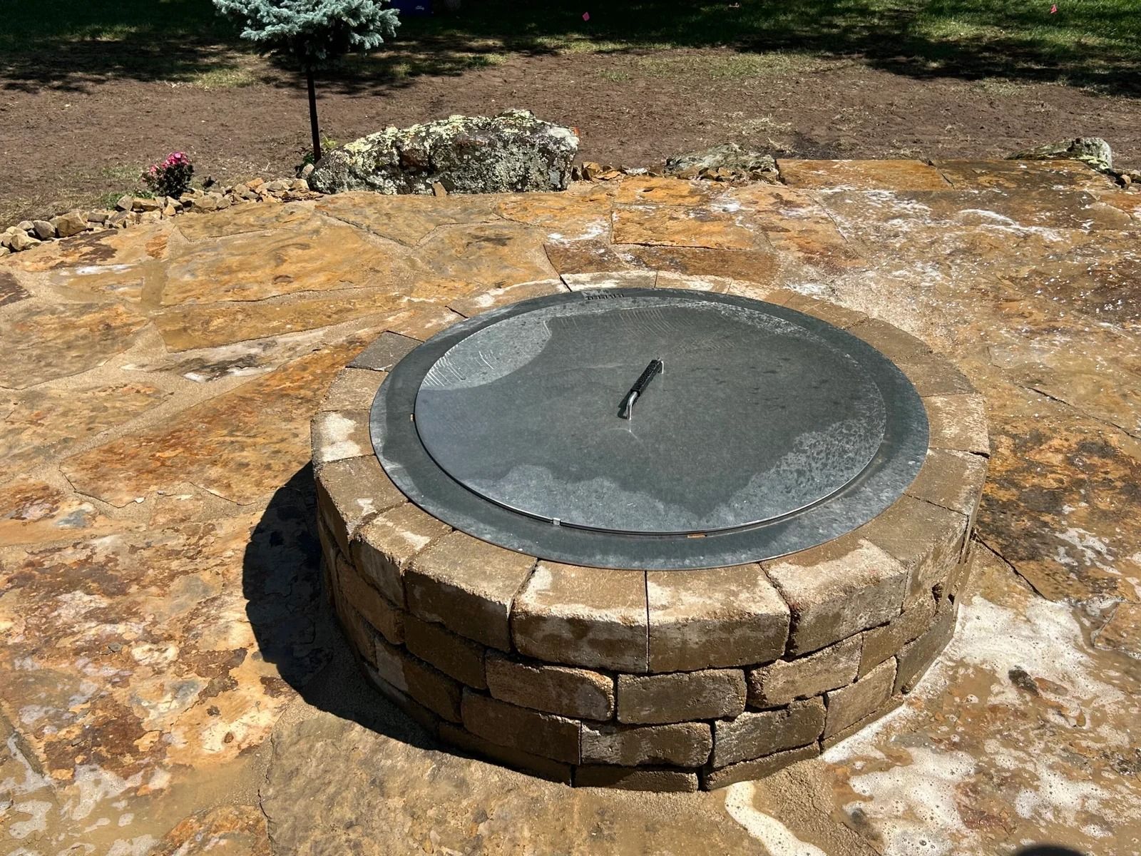 Round stone fire pit with a metal cover set in a rocky outdoor yard