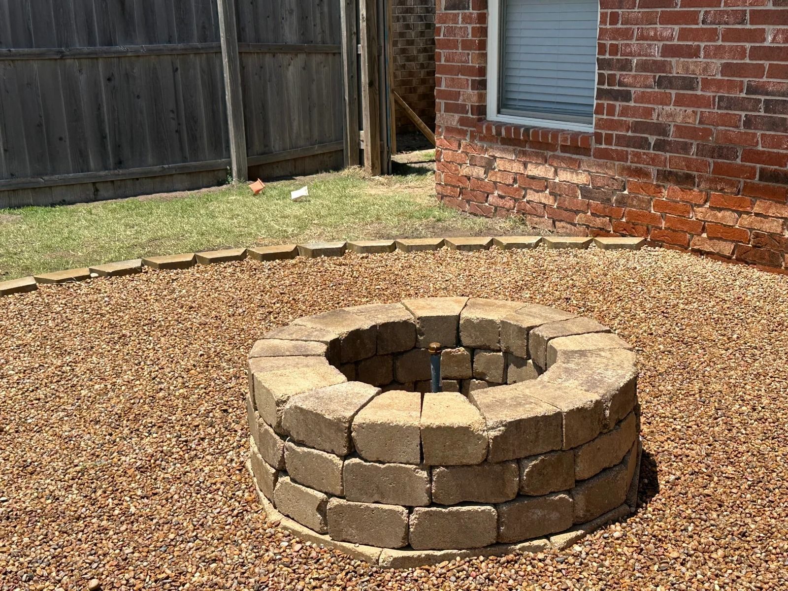 Stone fire pit in a gravel backyard next to a brick house and wooden fence.