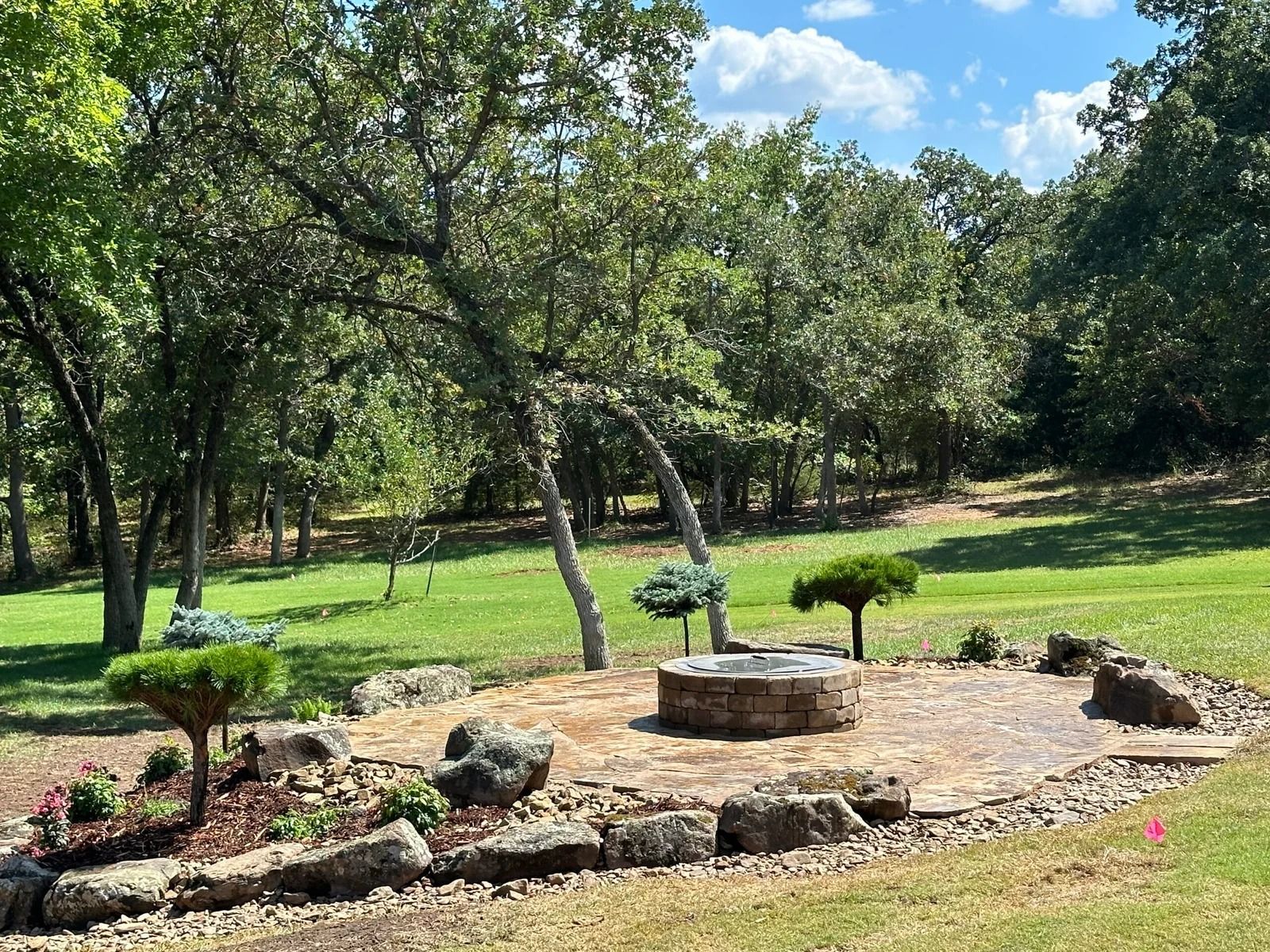 Landscaped garden with a stone fire pit, mulch beds, and trees under a sunny blue sky