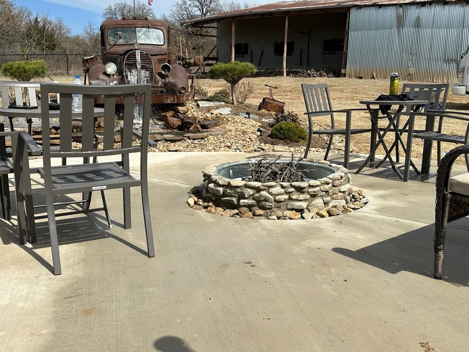 Rustic patio with metal chairs and table around a stone fire pit, old truck and barn in the background