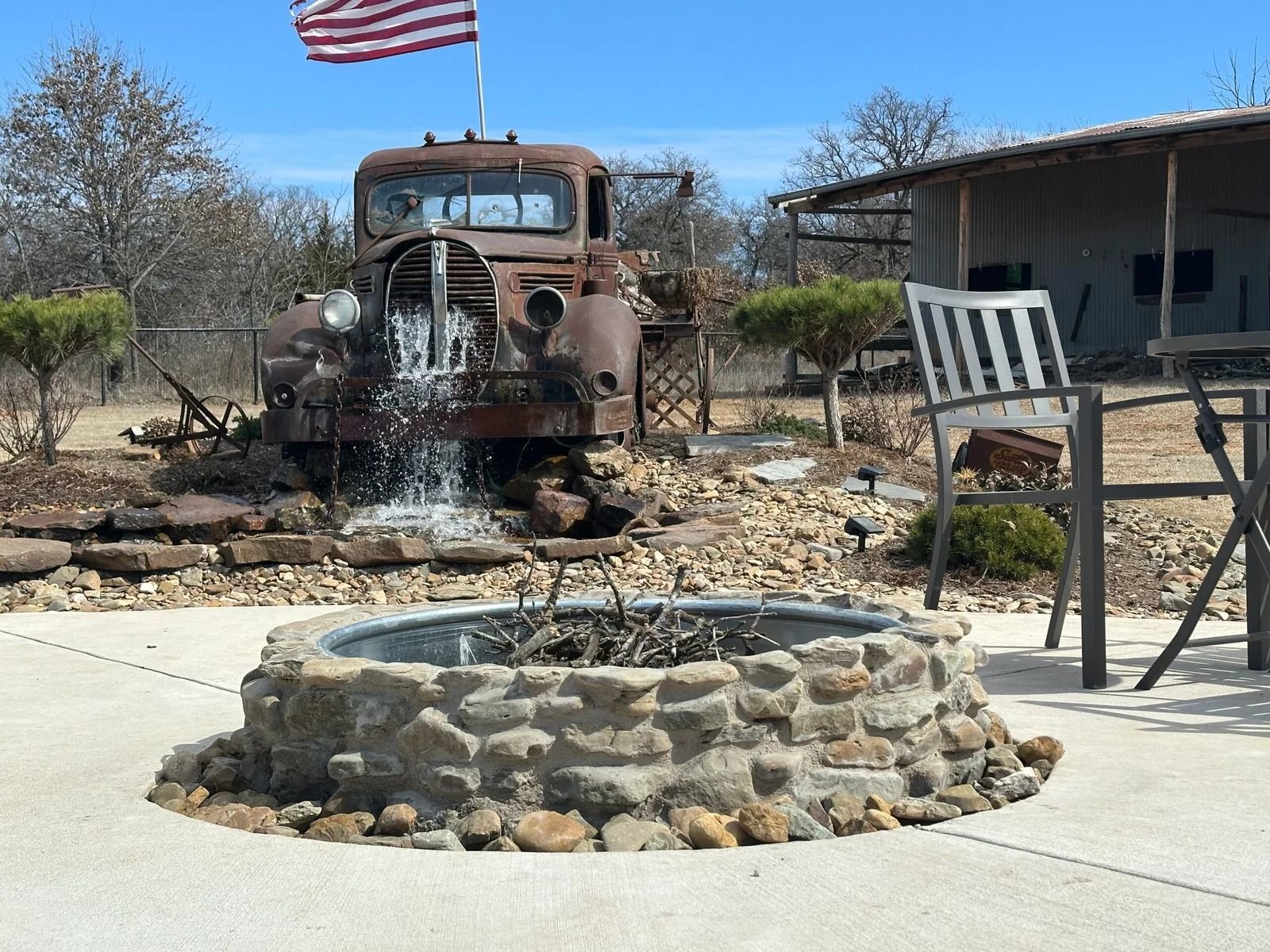 Rusty old truck behind a stone fire pit in a dry yard, with an American flag and patio chair nearby