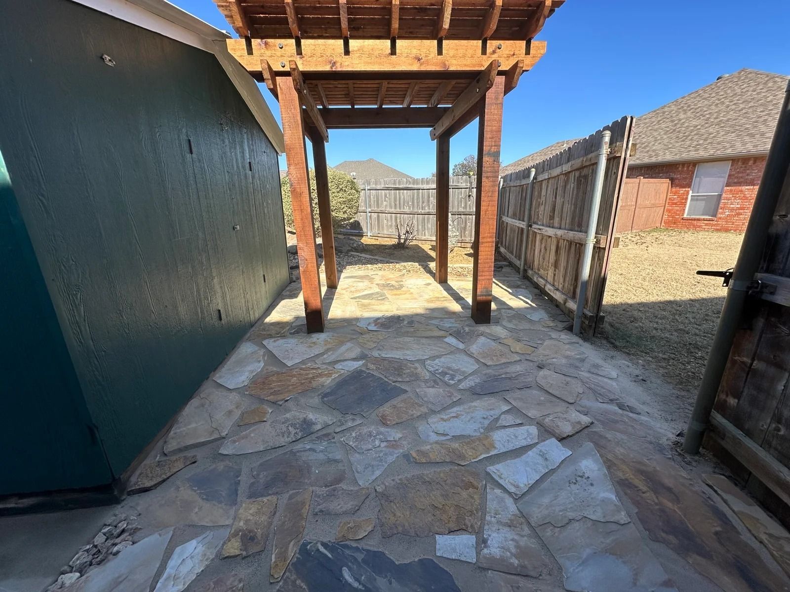 Stone patio walkway under a wooden pergola between a teal wall and a fence, with mountains in the distance