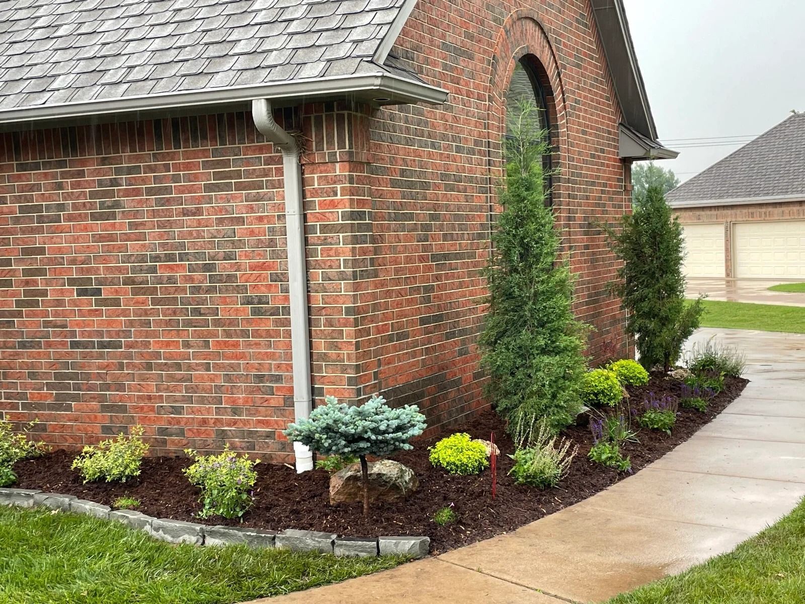 Brick house exterior with arched window, landscaped shrubs, and a curved concrete walkway