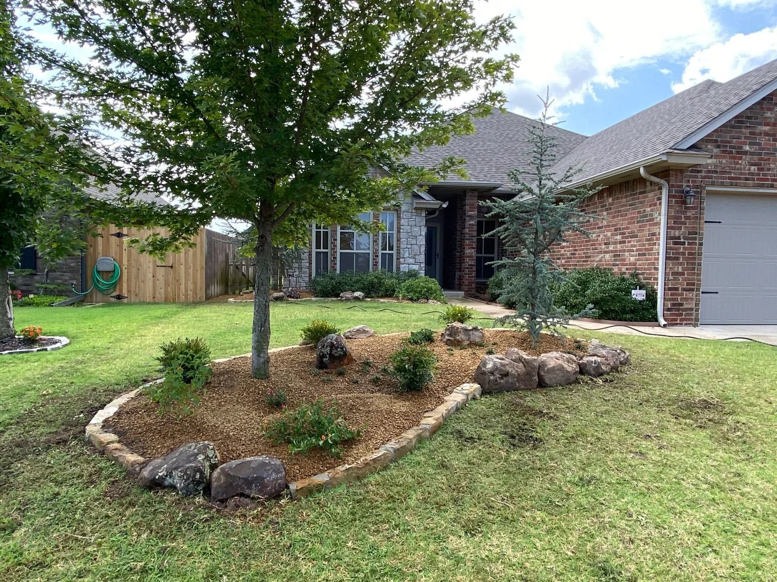 Front yard landscaping with a tree, stone border, mulch bed, and a brick house with a garage door.