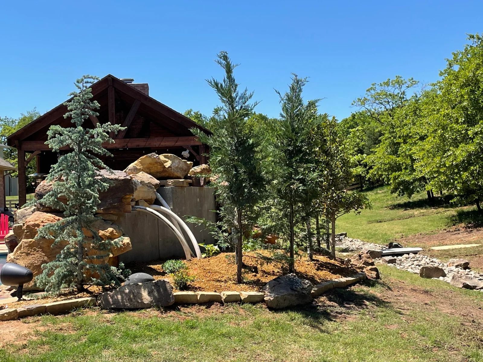 Backyard garden with a small wood gazebo, large rocks, trees, and a sunny blue sky