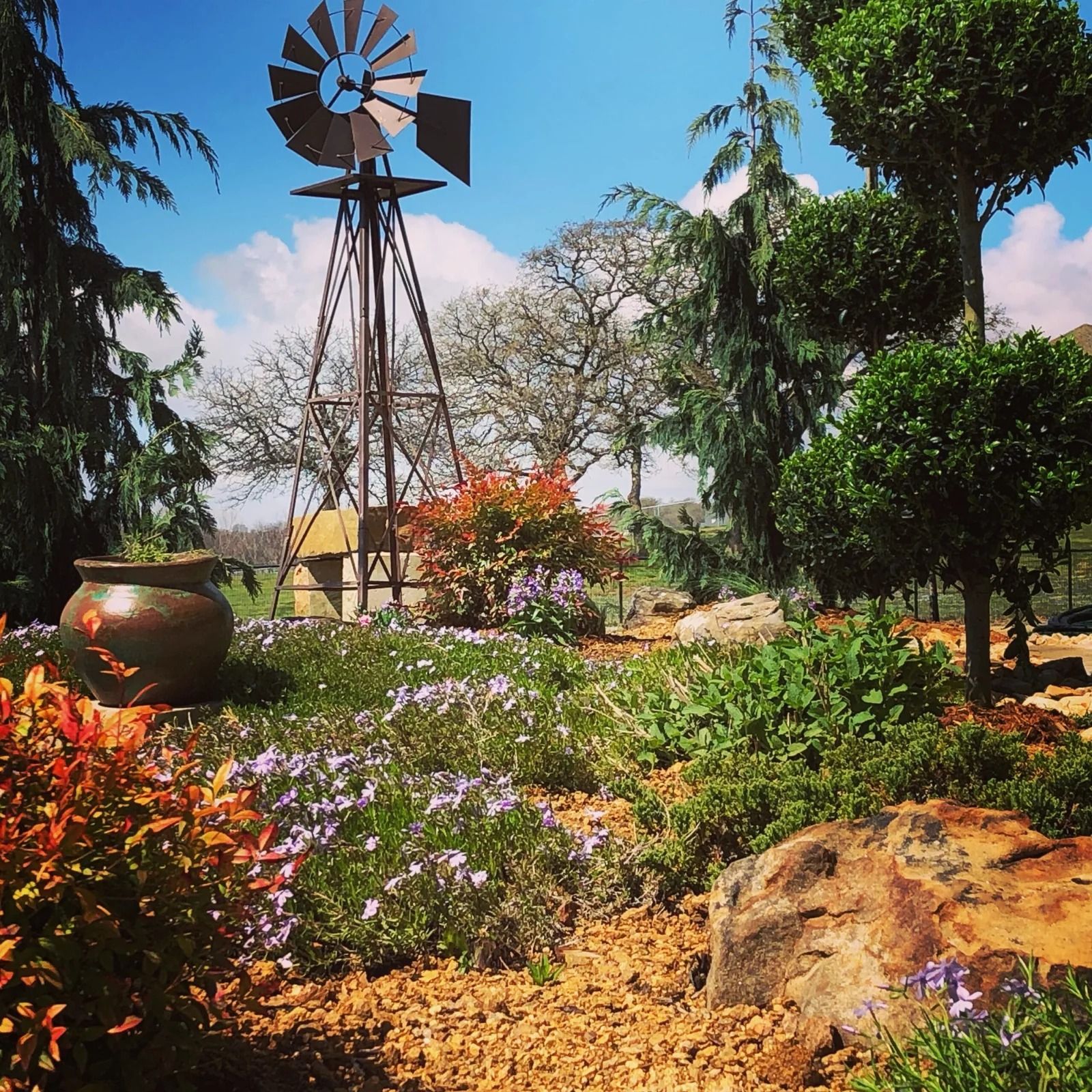 Windmill above a colorful garden with flowers, rocks, and trees under a bright blue sky