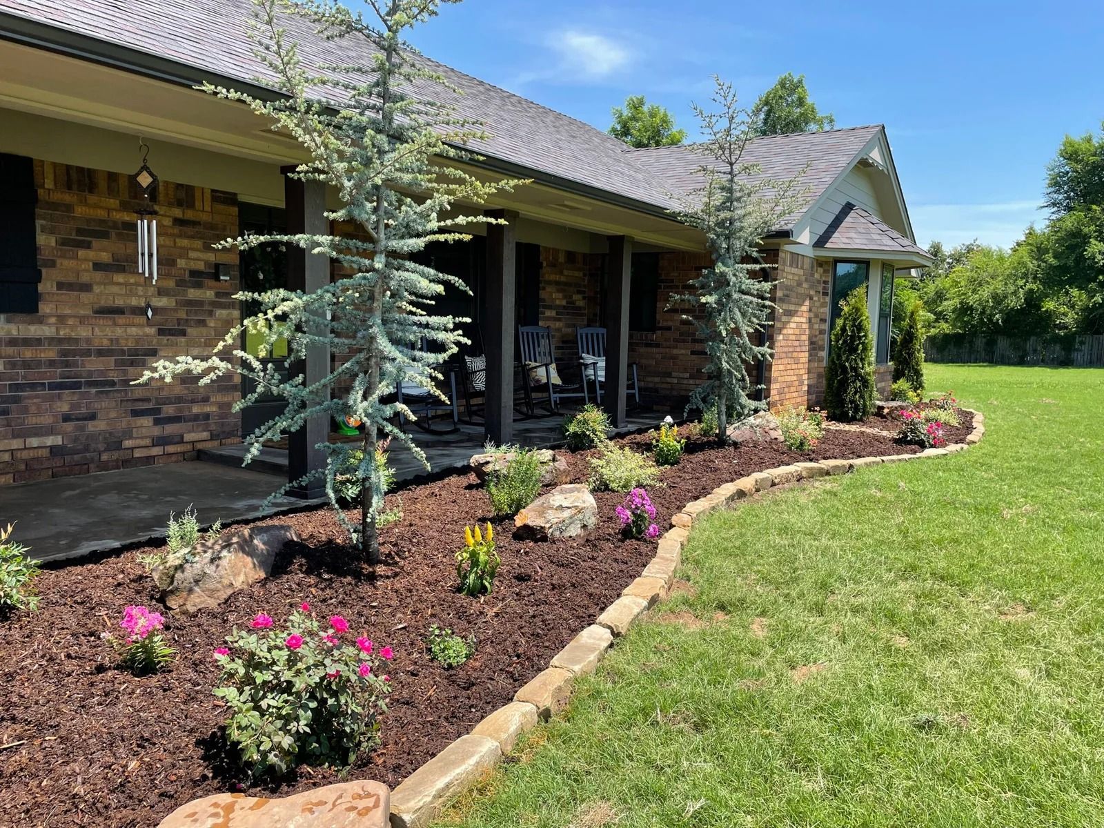 Brick house front yard with newly planted flower bed, small trees, and green lawn under a sunny blue sky