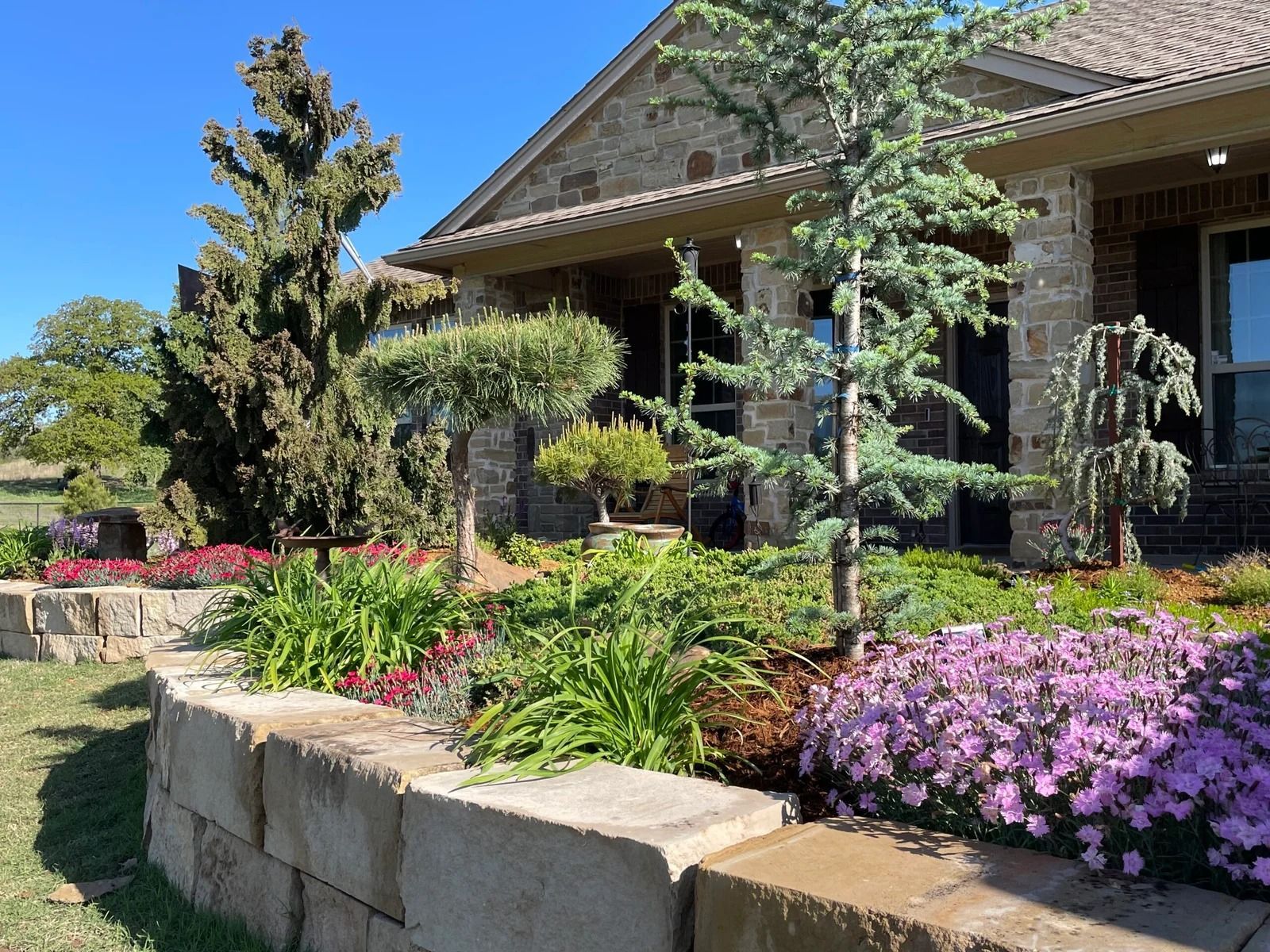 Landscaped garden with pink flowers and a stone retaining wall beside a house porch