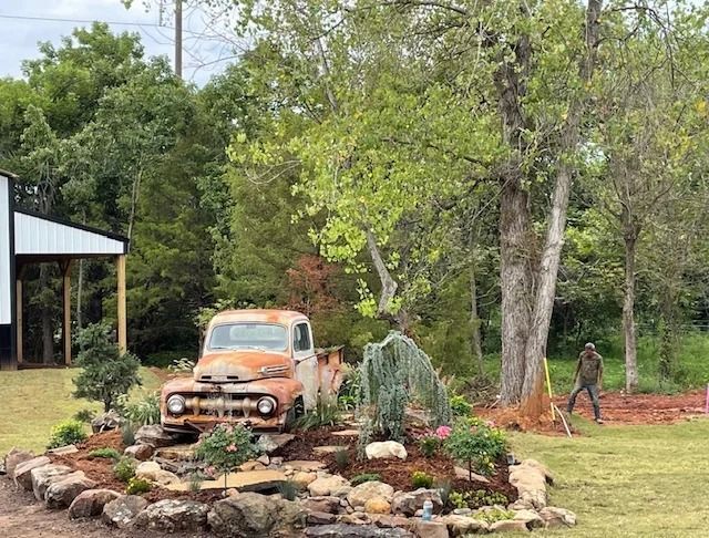 Old rusty pickup truck in a landscaped garden with rocks, trees, and a shed nearby